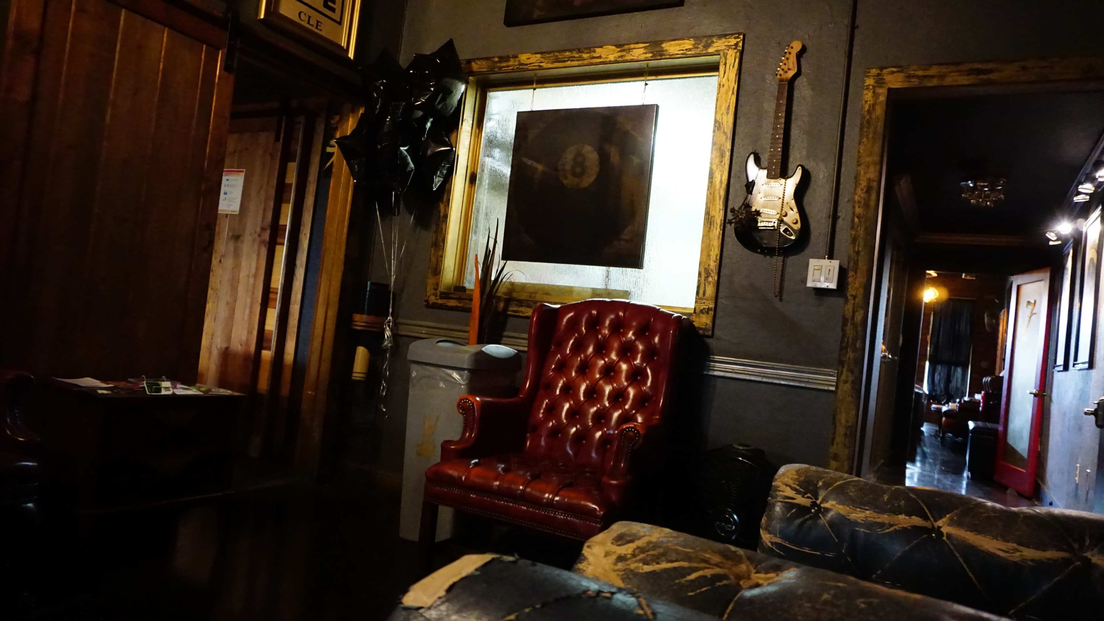 The image shows a dimly lit room featuring a vintage red chair, a guitar hanging on the wall, and dark, worn furniture.