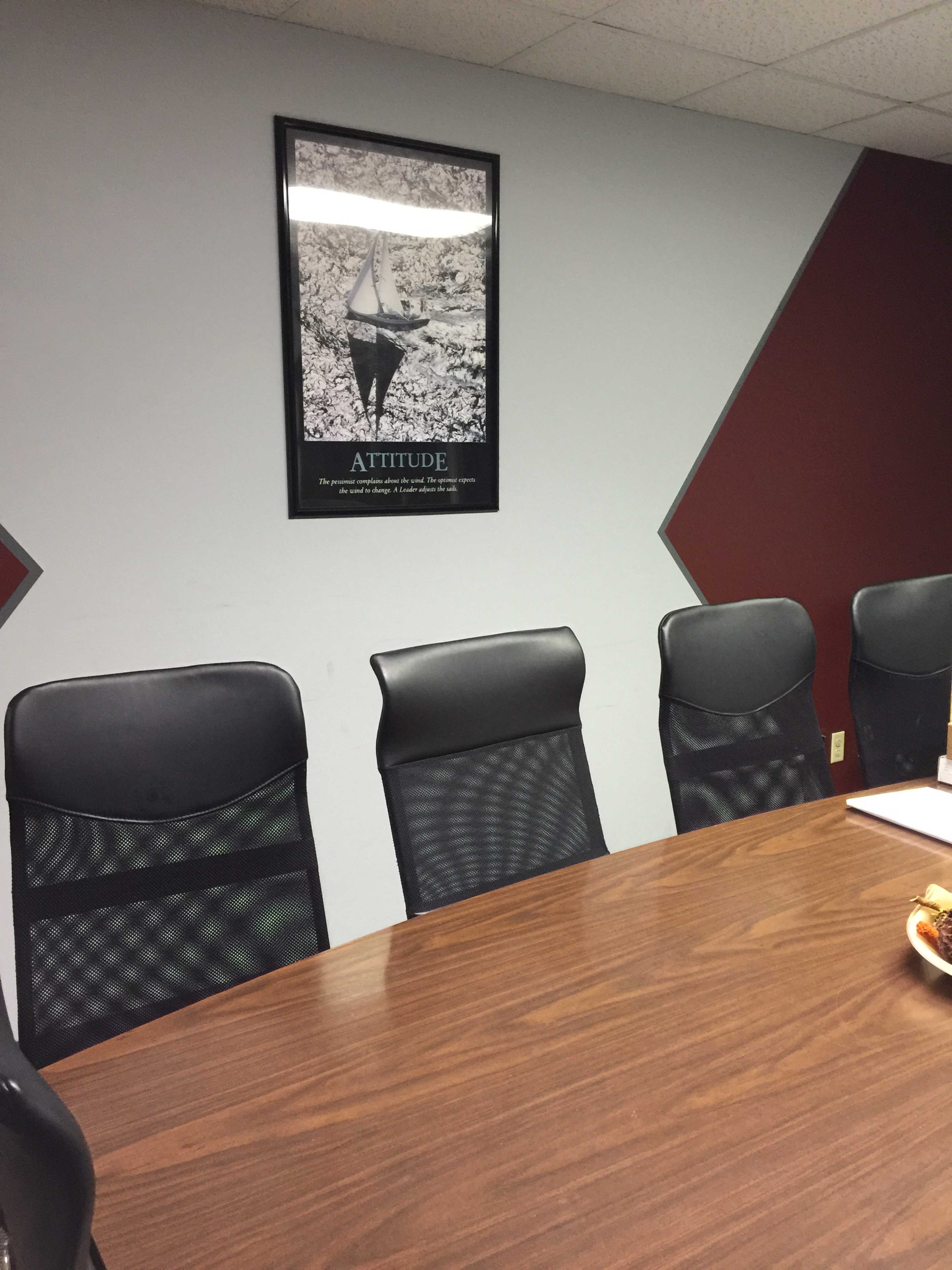 A conference room features a round wooden table surrounded by several black chairs, with a motivational poster titled "ATTITUDE" hanging on the wall.