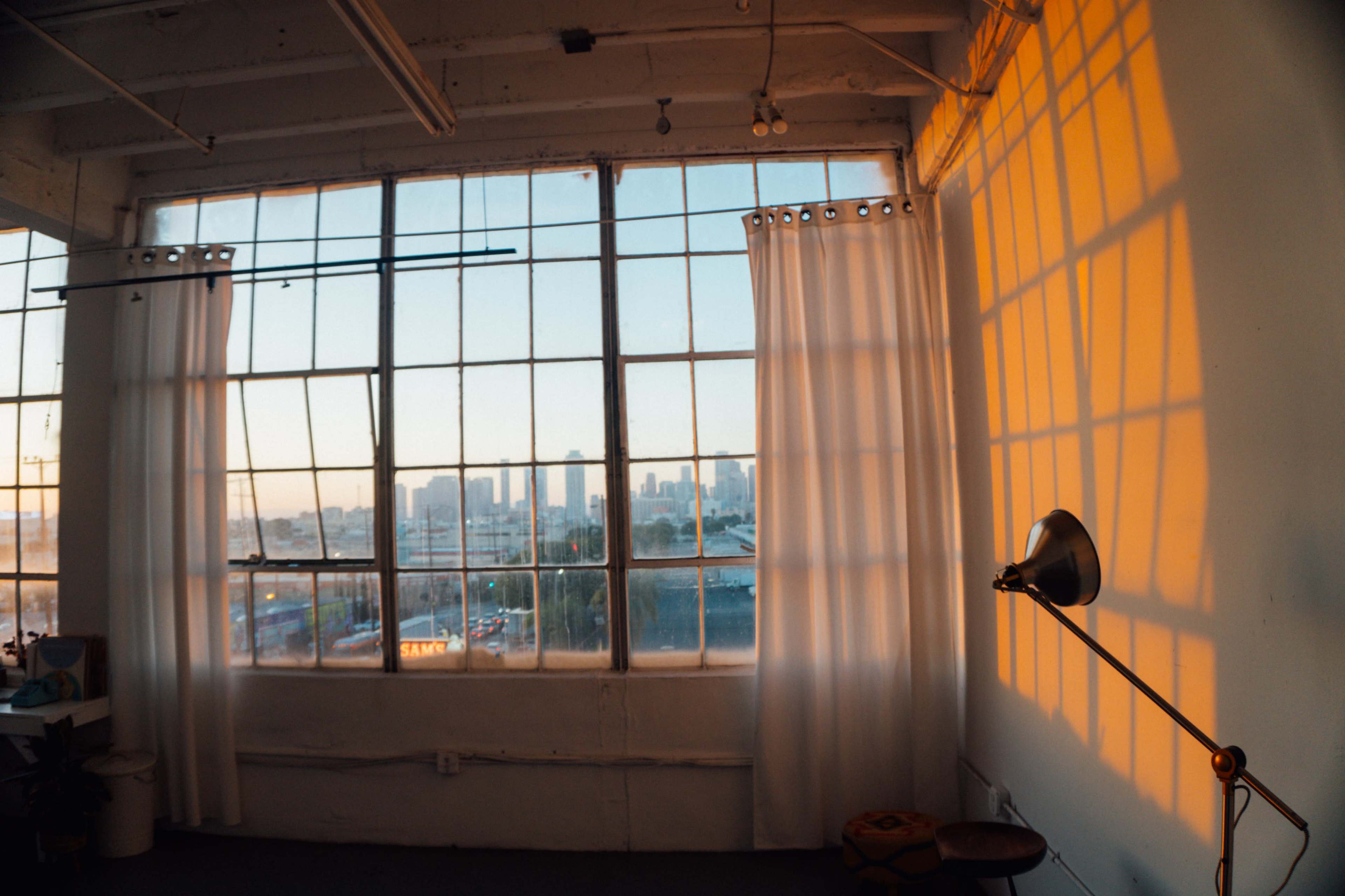 The image shows a spacious room with large windows allowing sunlight to cast shadows on the wall, revealing a city skyline in the background.