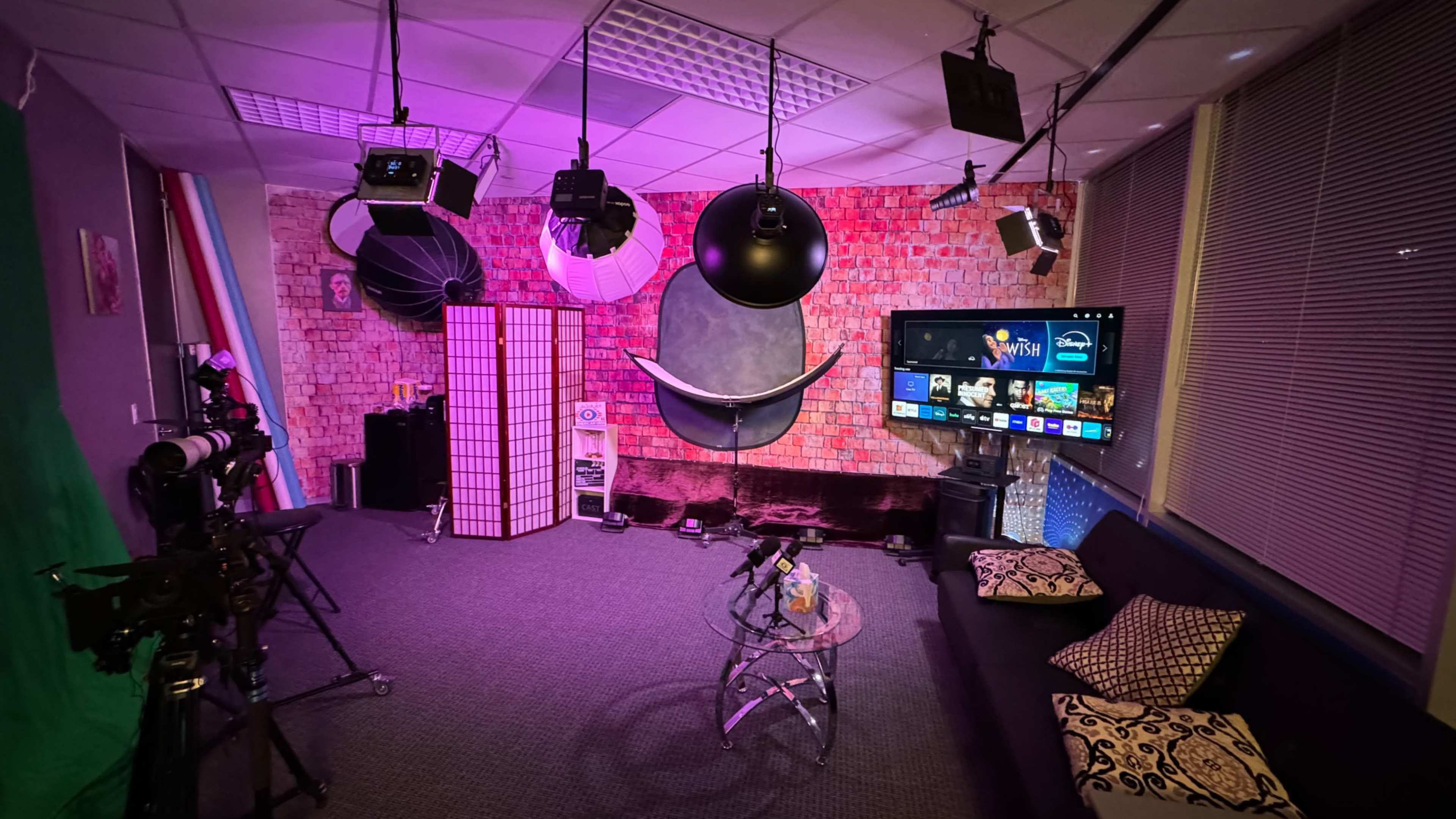 A studio setup with a black sofa, a glass coffee table, a television screen, and various lighting equipment against a brick wall backdrop.