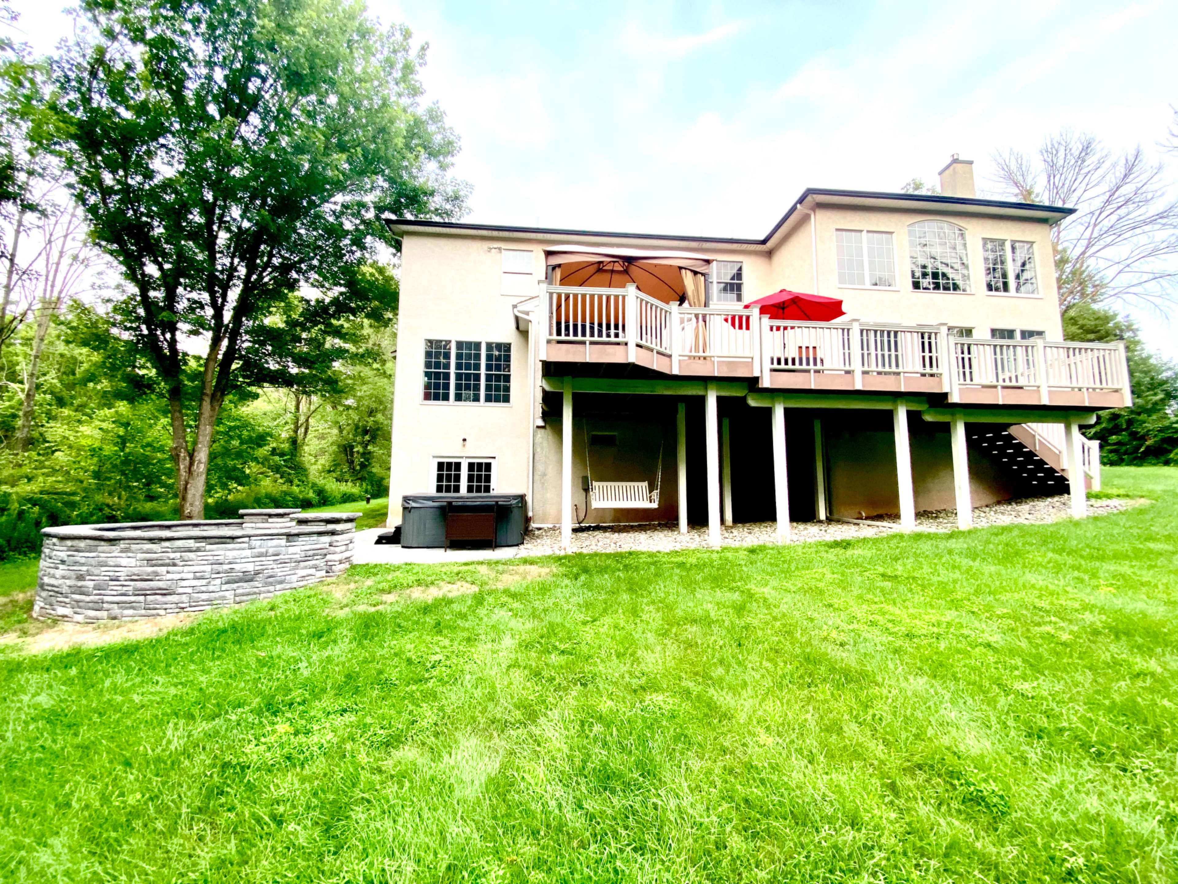 The image shows a two-story house with a deck and patio furniture, surrounded by a grassy yard and a stone fire pit.