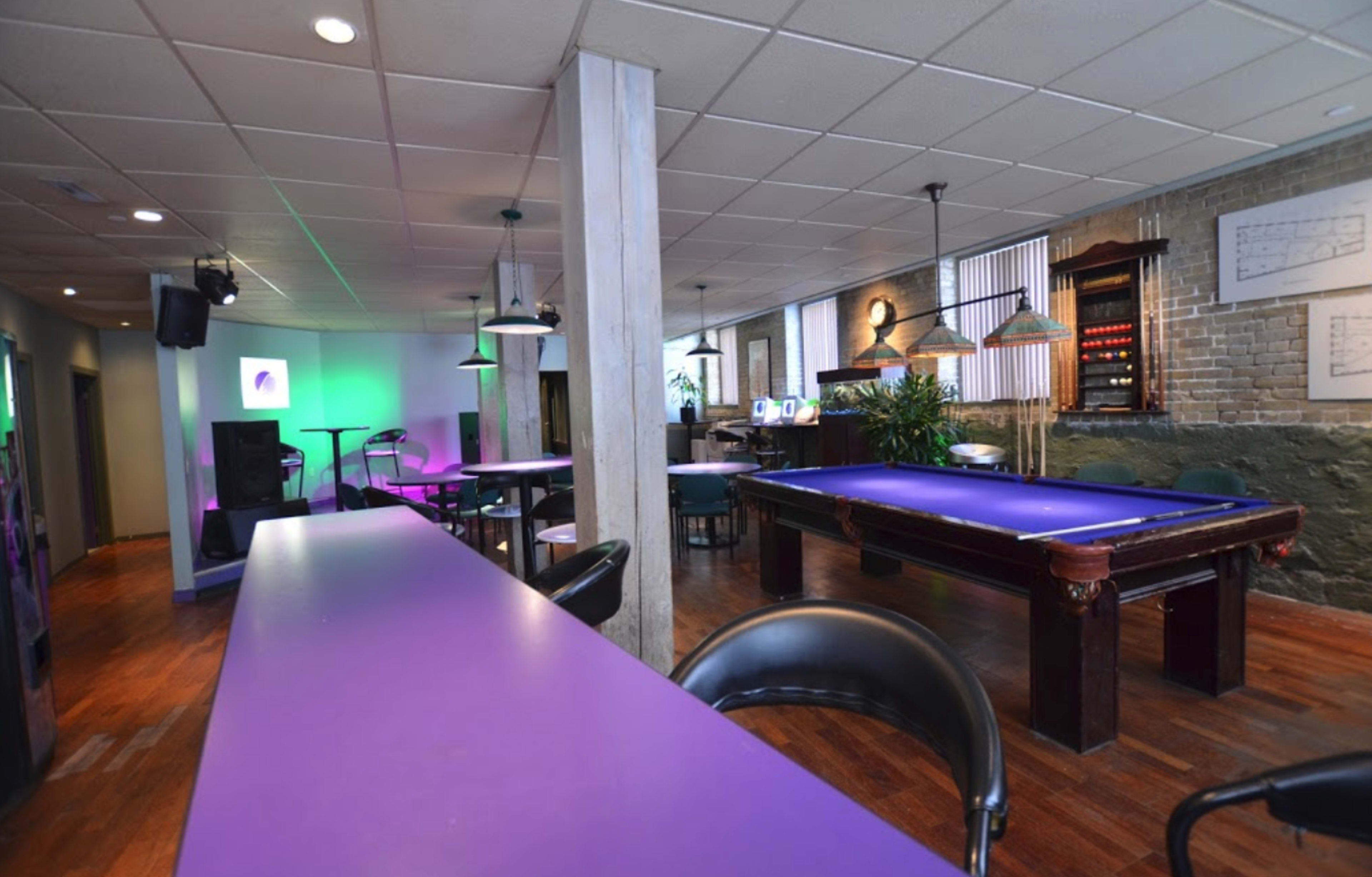 The image shows a spacious room featuring a purple pool table, bar counter, and seating areas with ambient lighting.