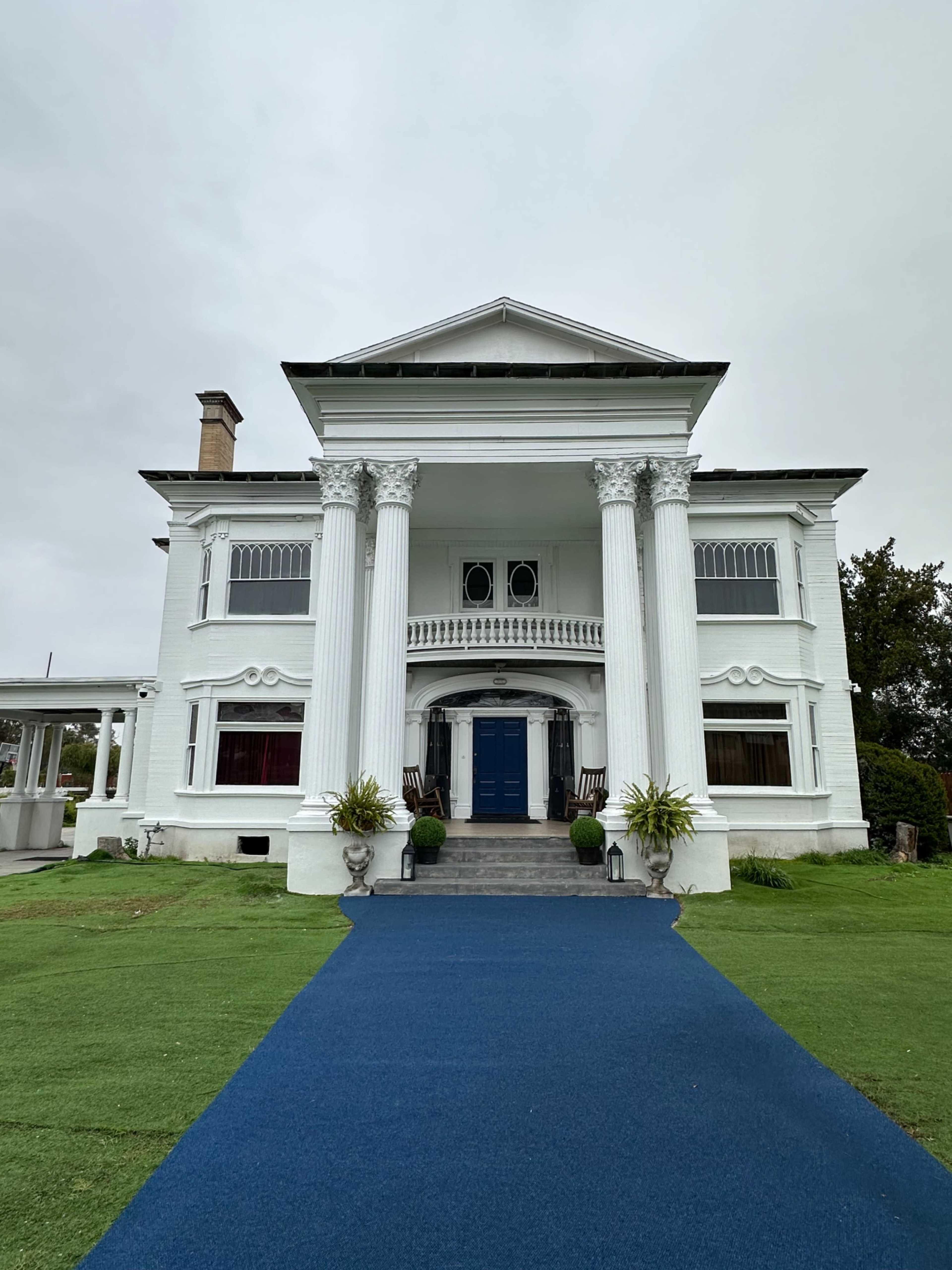 The image shows a large white columned mansion with a blue pathway leading to the front door.