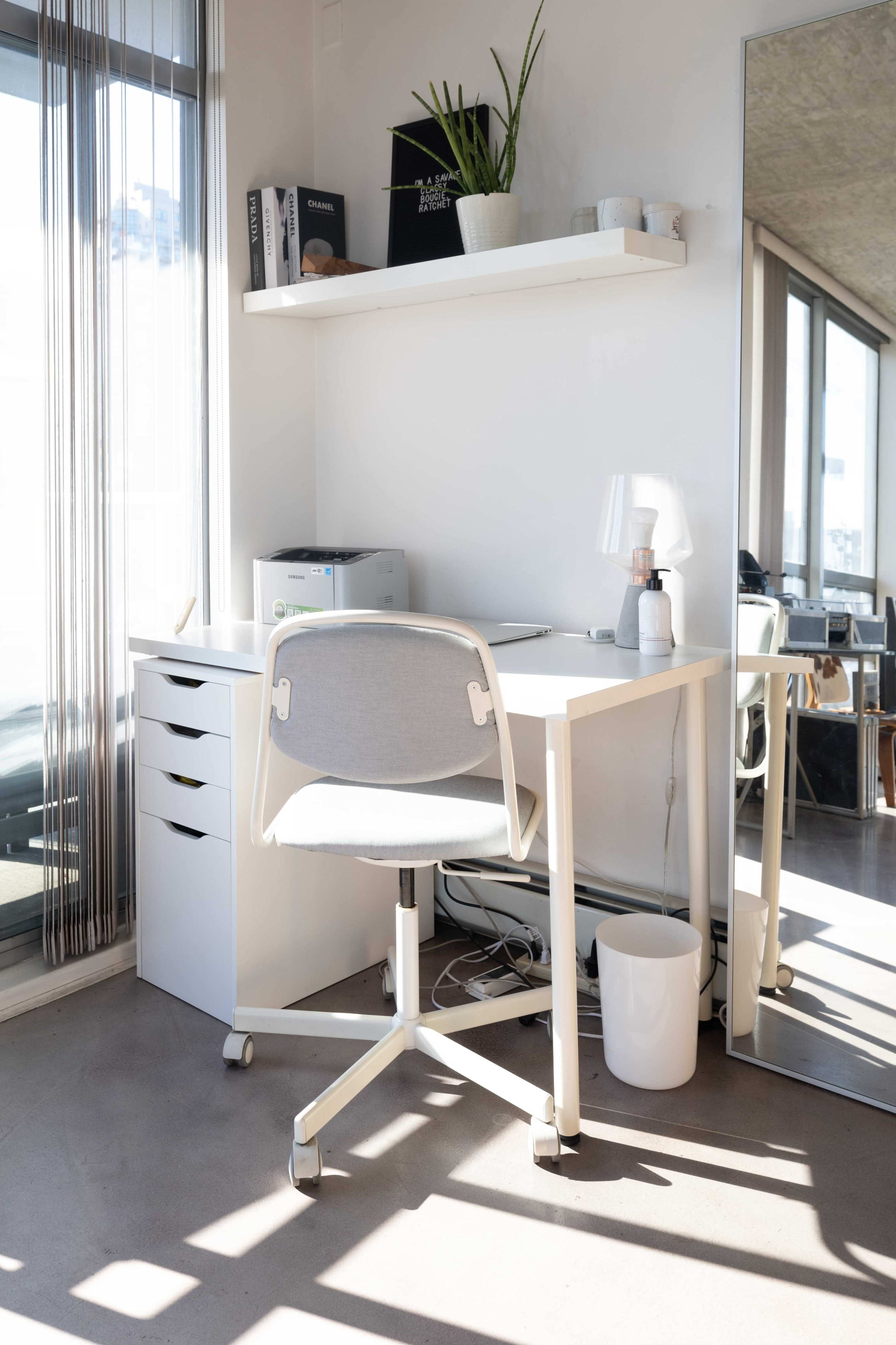 A modern workspace features a white desk with a chair, a shelf above, a printer, and large windows letting in natural light.