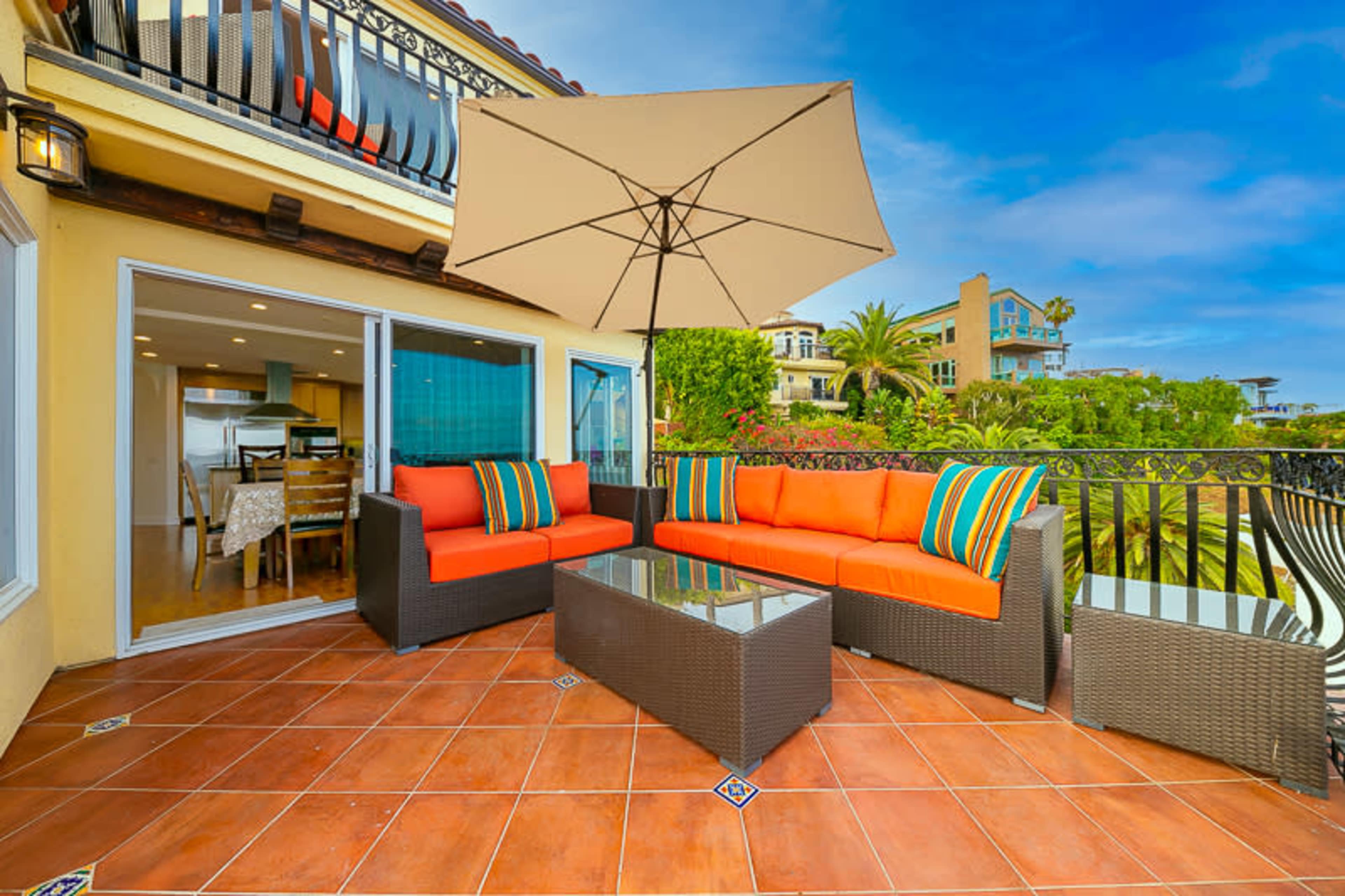 A patio features orange seating and a glass-topped table under a large umbrella, overlooking a neighborhood with palm trees.