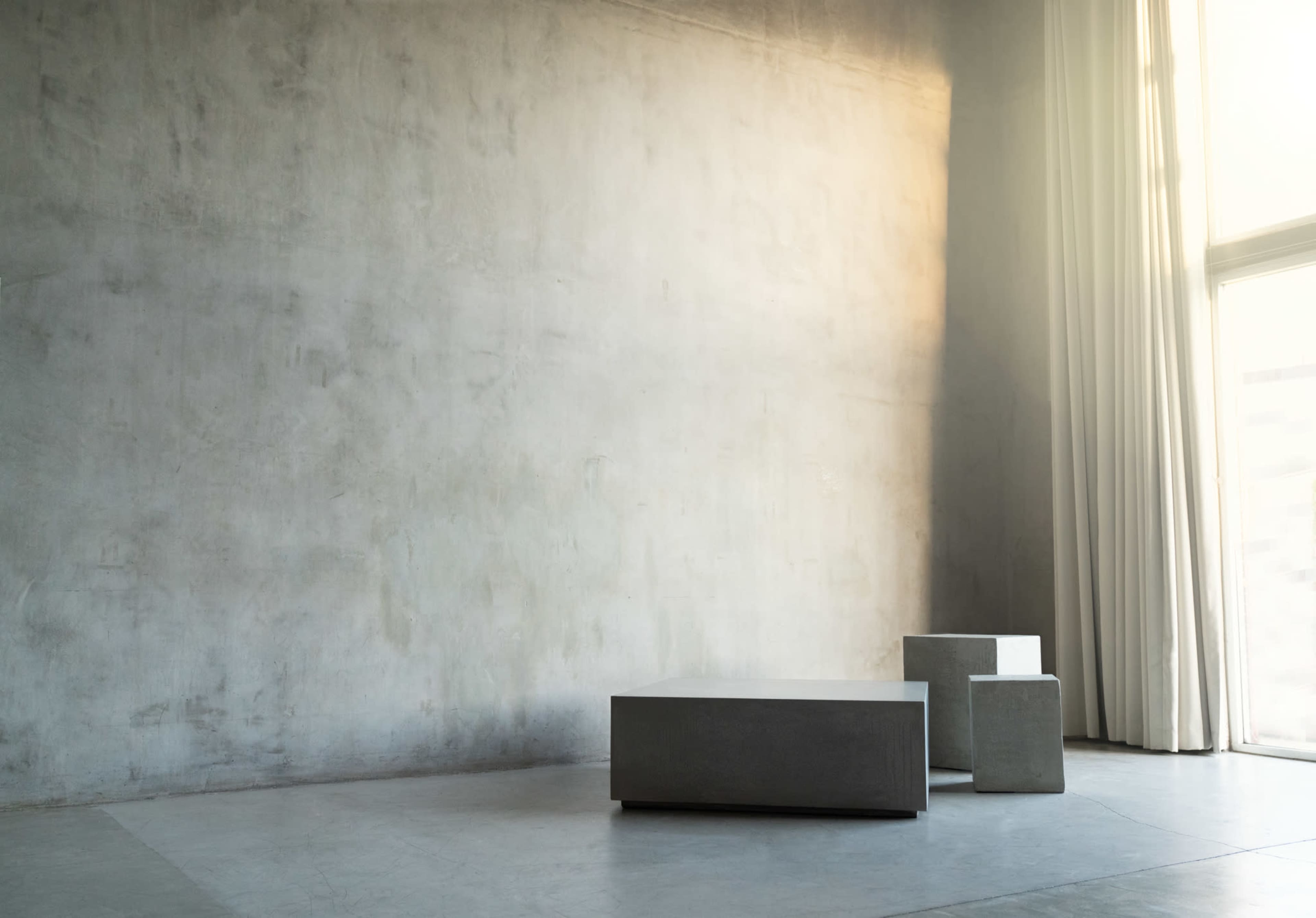 The image shows a minimalistic interior space with a textured gray wall, two geometric concrete tables, and large windows covered by sheer curtains.