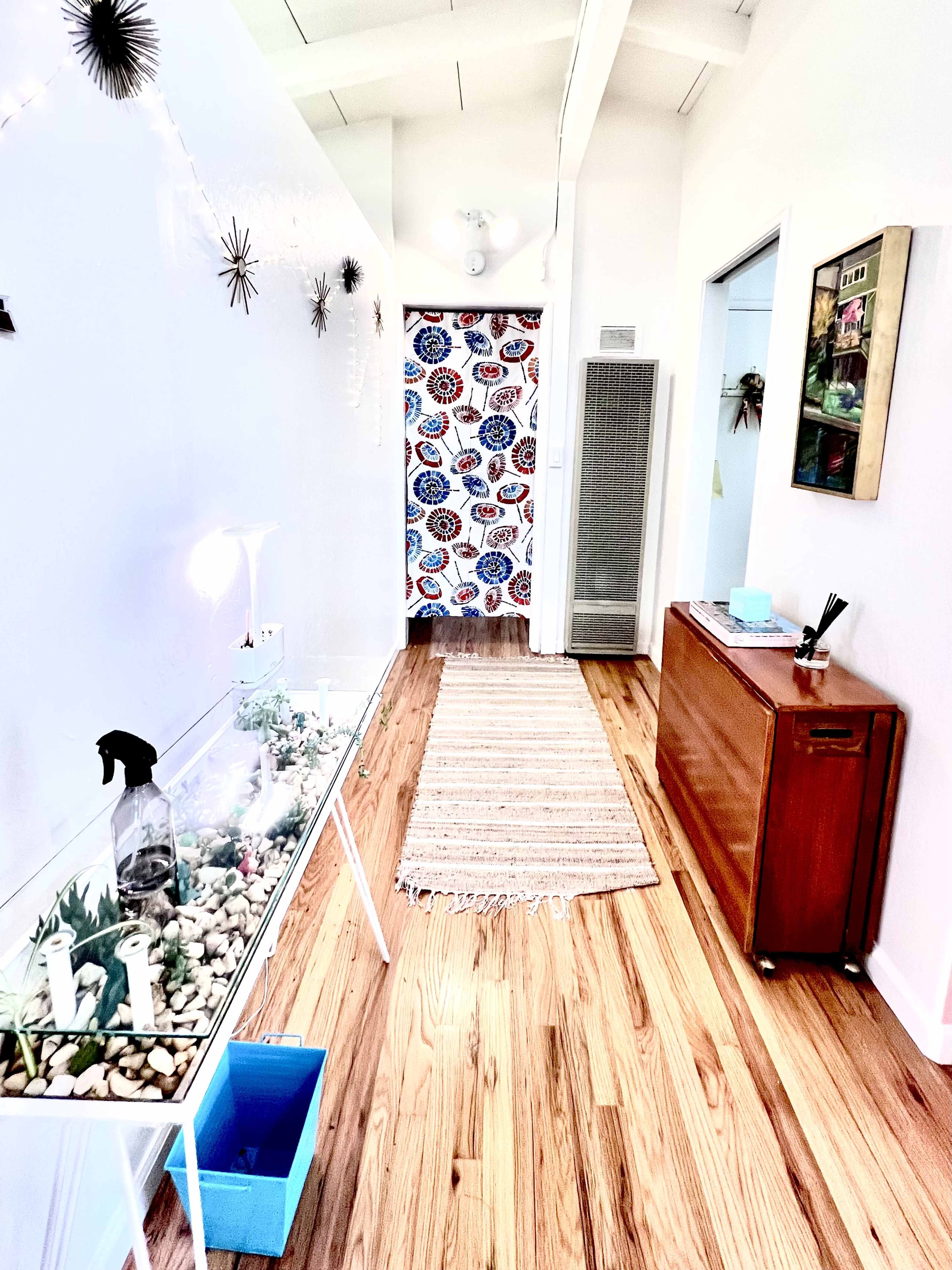 A hallway features a glass table with a decorative arrangement, a colorful patterned door at the end, and wooden flooring.