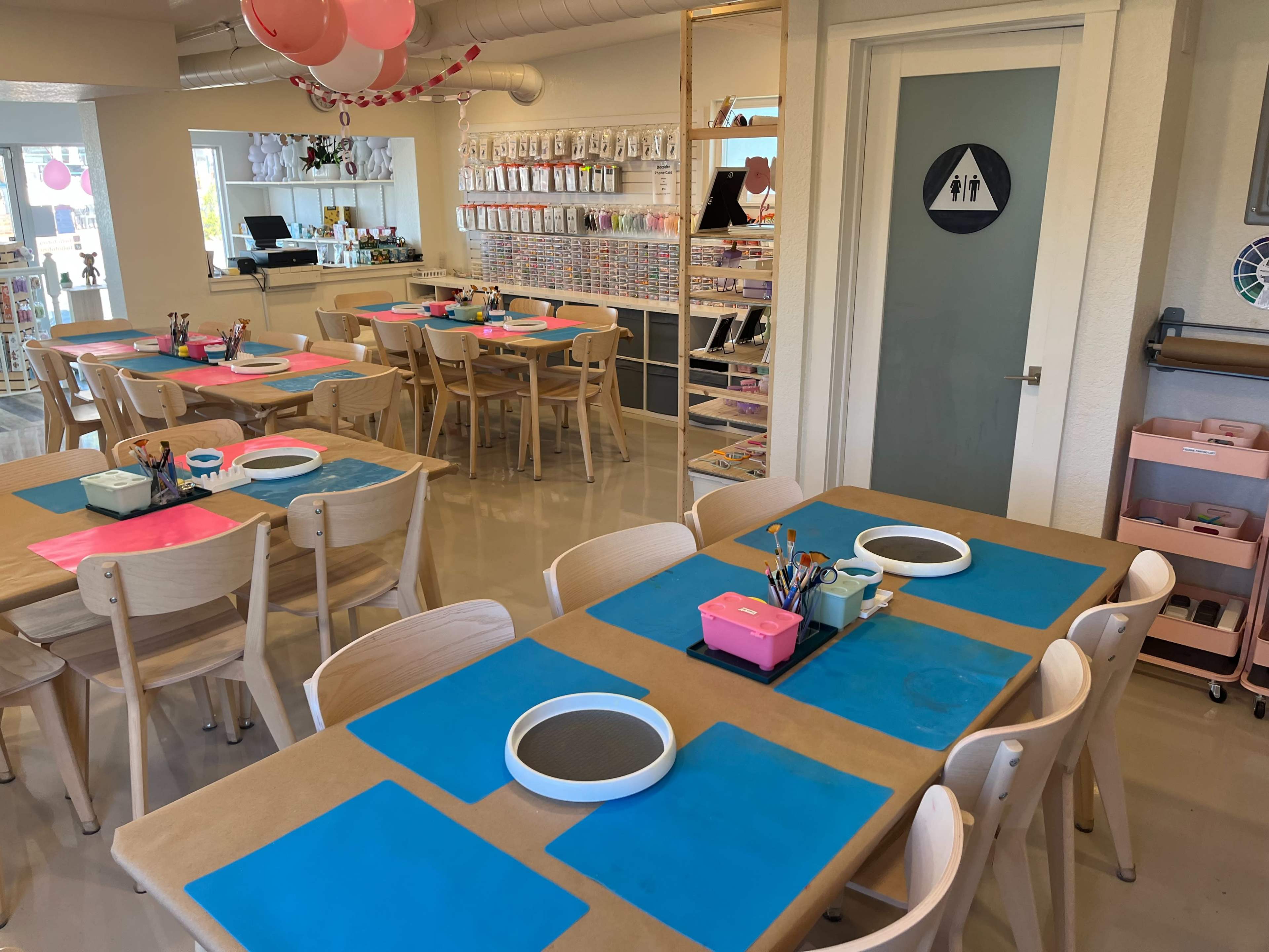 The image shows a bright, organized art studio with tables covered in blue and pink paper, surrounded by wooden chairs and craft supplies.