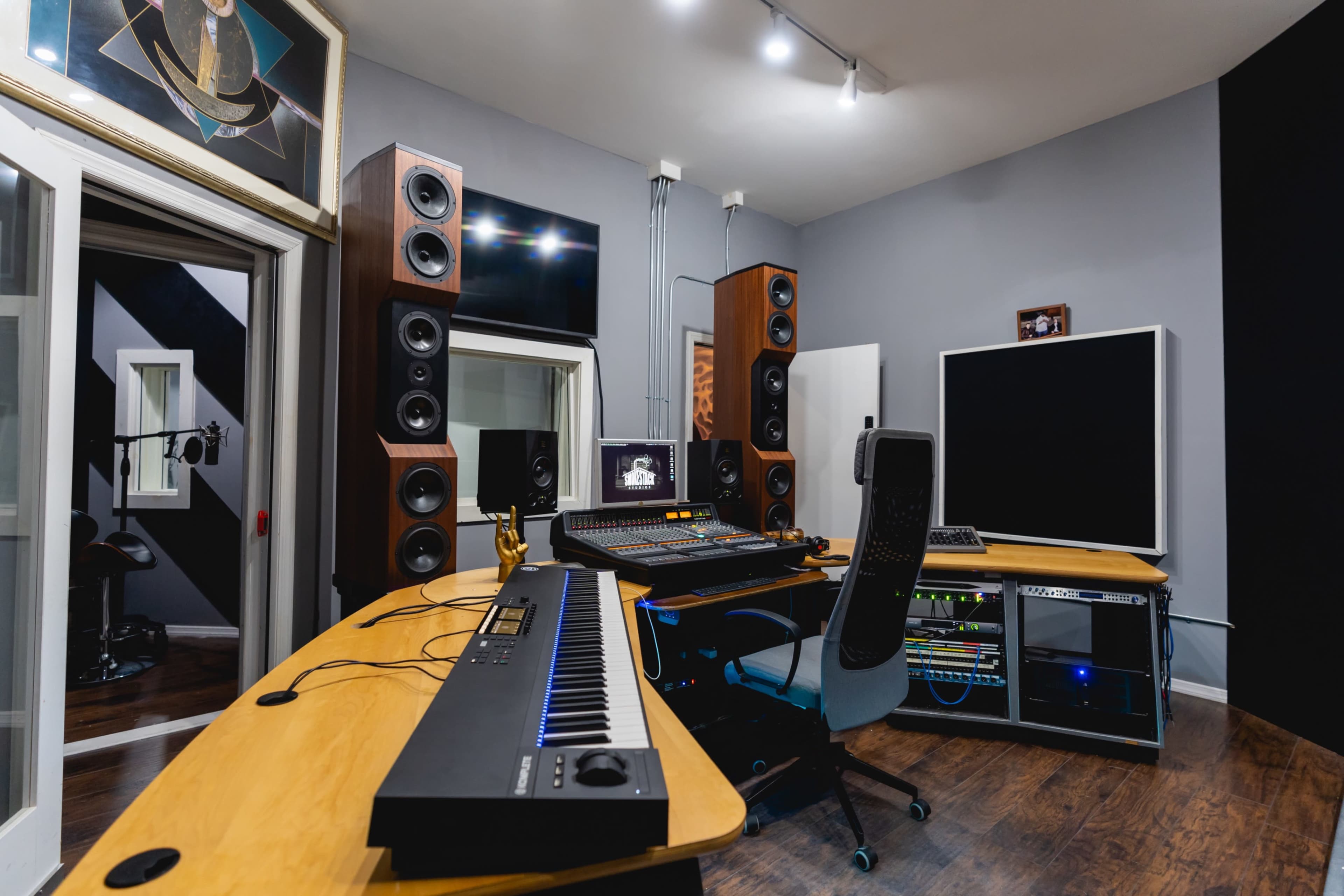 The image shows a modern music studio with a large desk containing a keyboard, audio equipment, and tall speakers, along with a sound booth and acoustic treatment on the walls.