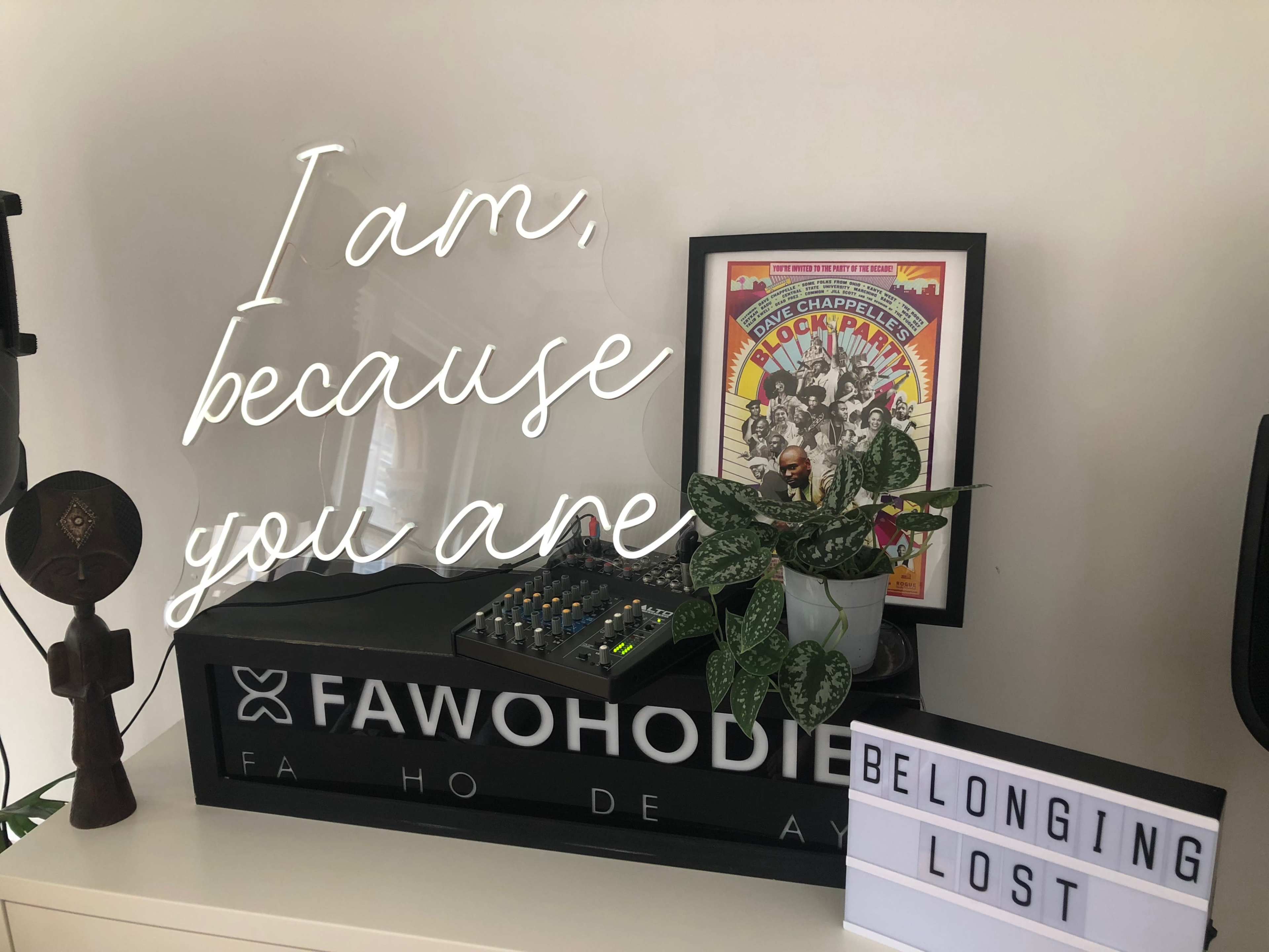 A modern desk displays a sound mixing console, a decorative plant, and lightbox signs with the words "I am, because you are" and "BELONGING LOST."