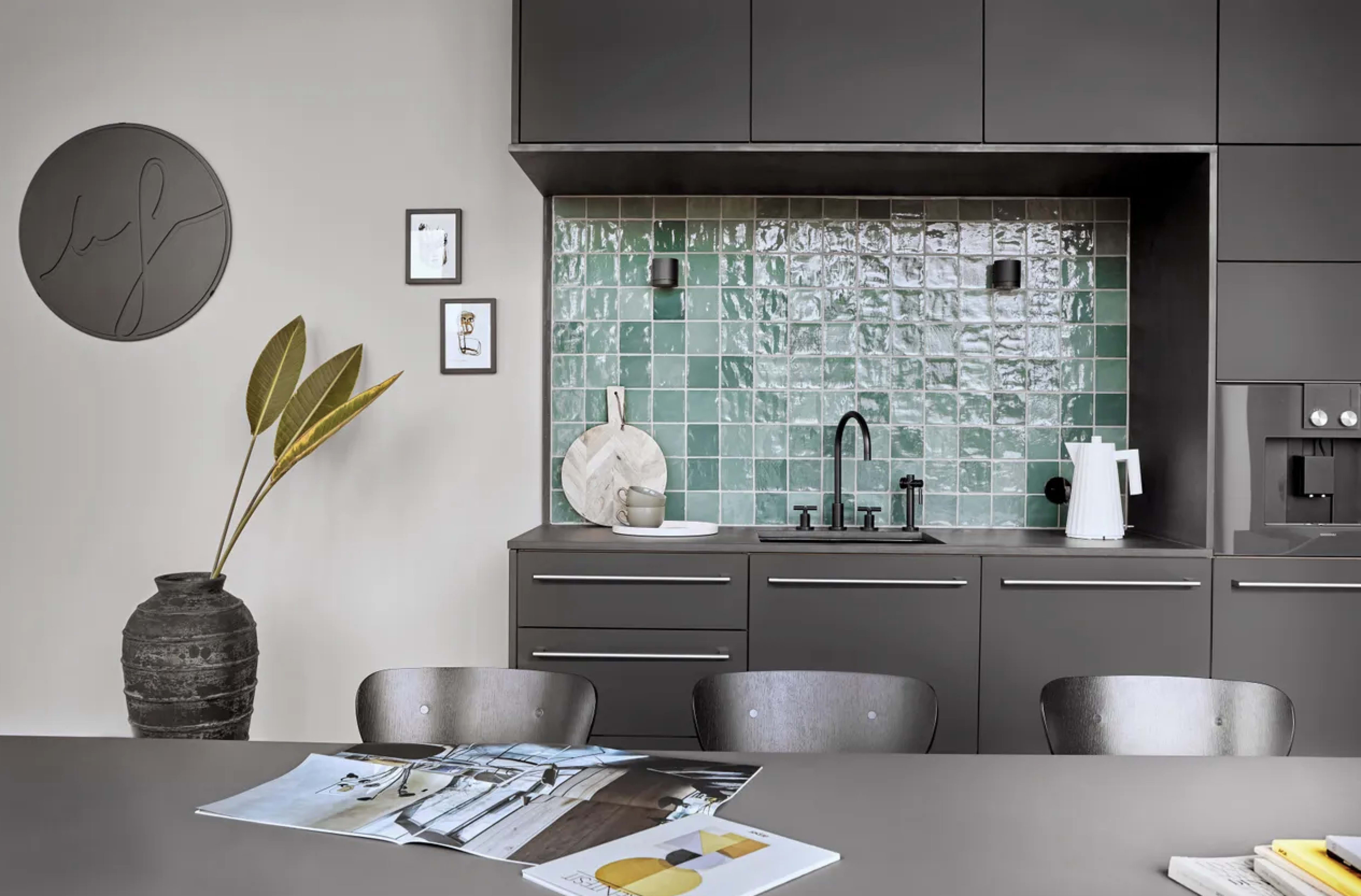 A modern kitchen features gray cabinetry, green tile backsplash, and a minimalist dining area with a plant and magazines on the table.