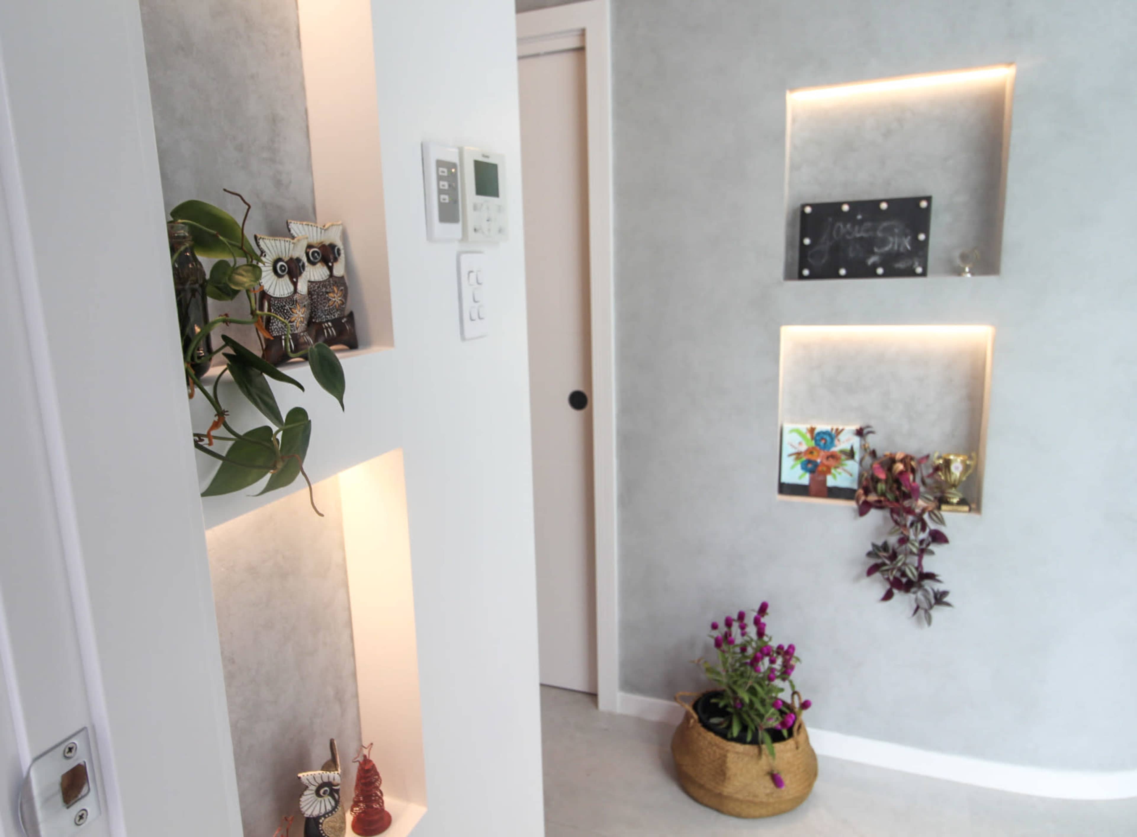 The image shows a modern interior hallway with recessed shelf lighting displaying decorative items and plants against a gray wall.