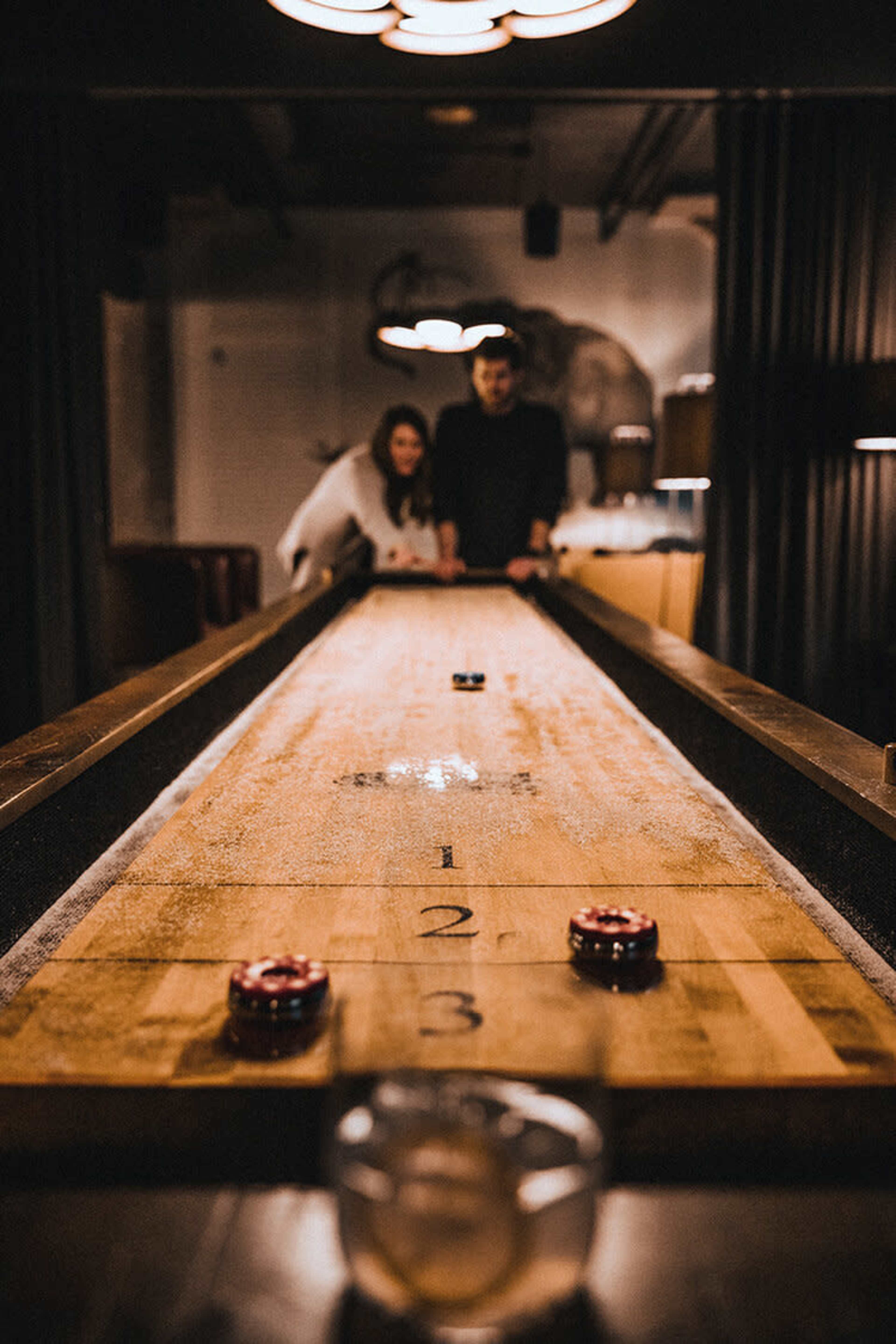 A wooden shuffleboard table is positioned in a dimly lit bar, with two people leaning over it, focusing on their game.