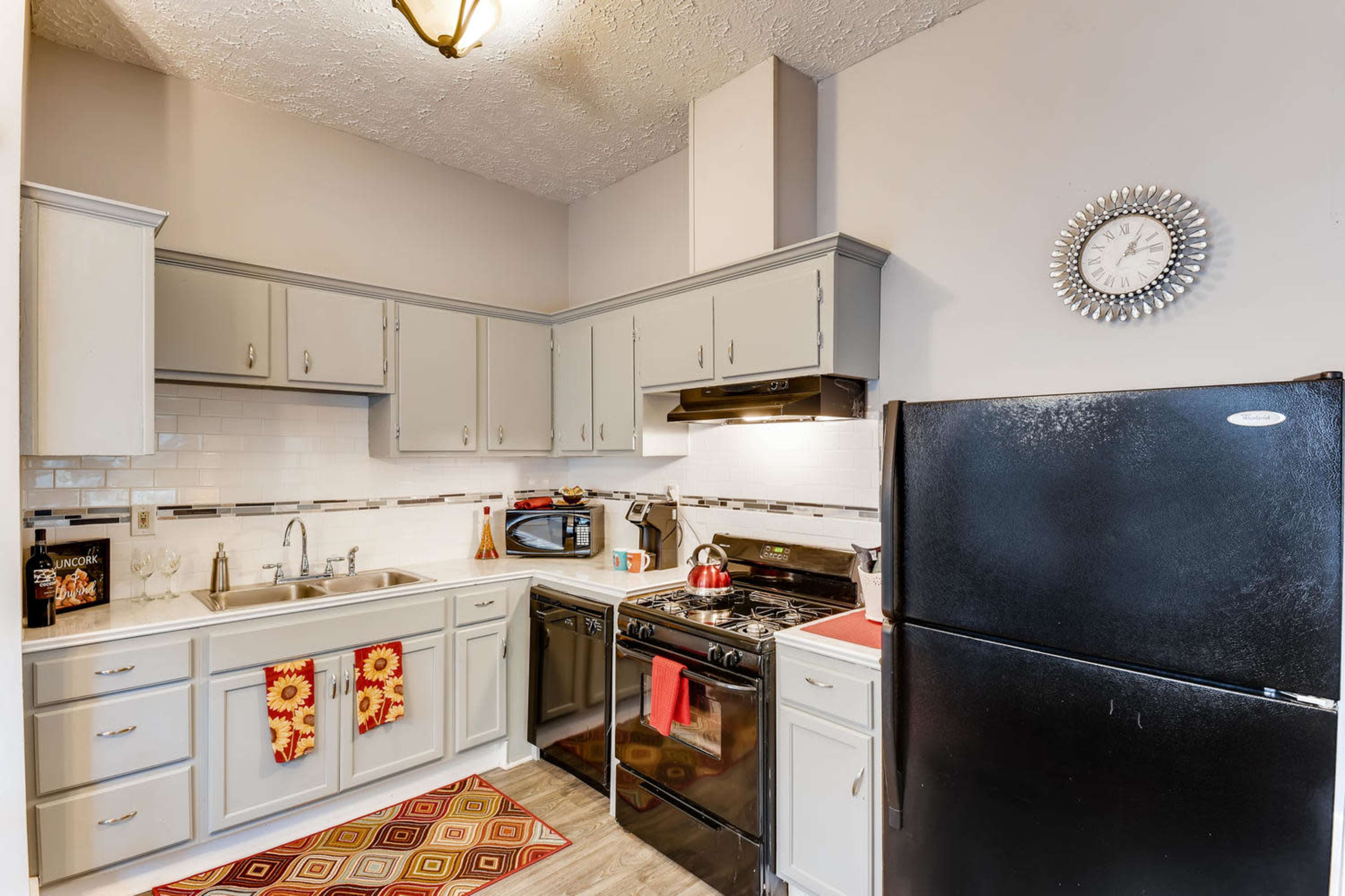 A compact kitchen features gray cabinetry, a black refrigerator, a stove with an oven, and a window above the sink adorned with a clock and decorative items.