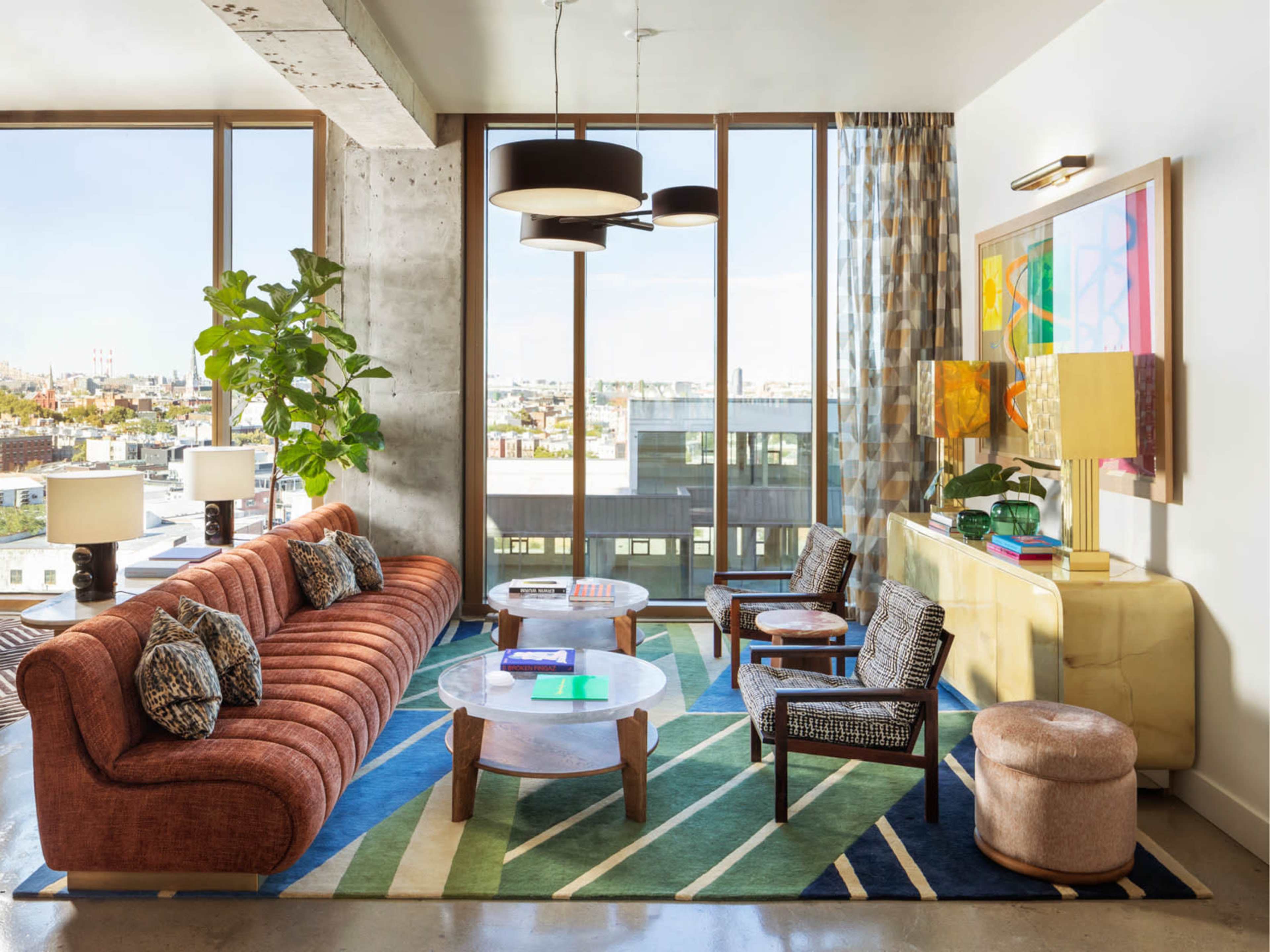 The image shows a modern living room with a large, curved sofa, a round coffee table, and a colorful rug, all set against large windows that provide a view of the city.