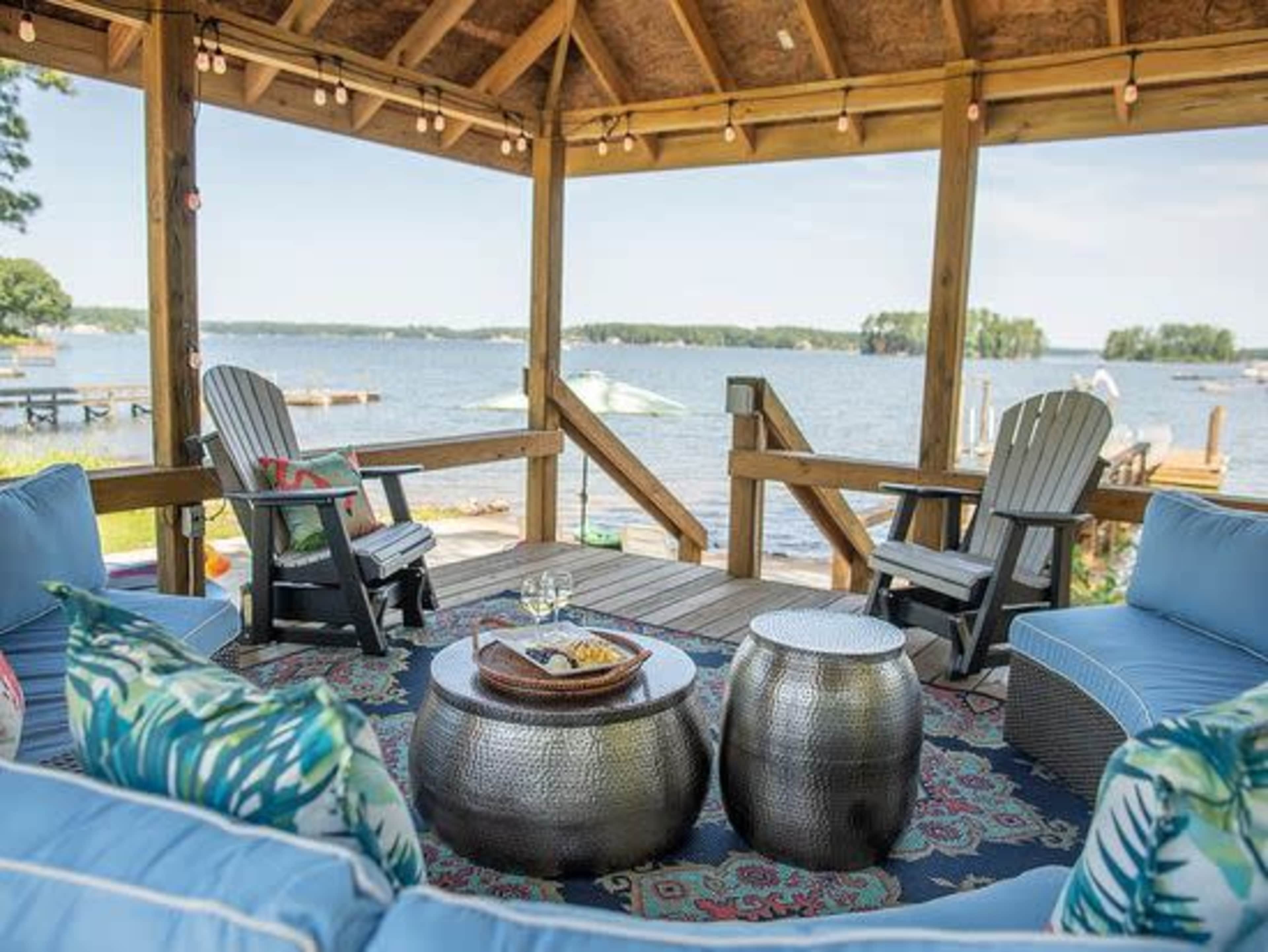 A covered wooden deck overlooking a lake features comfortable seating and decorative side tables.
