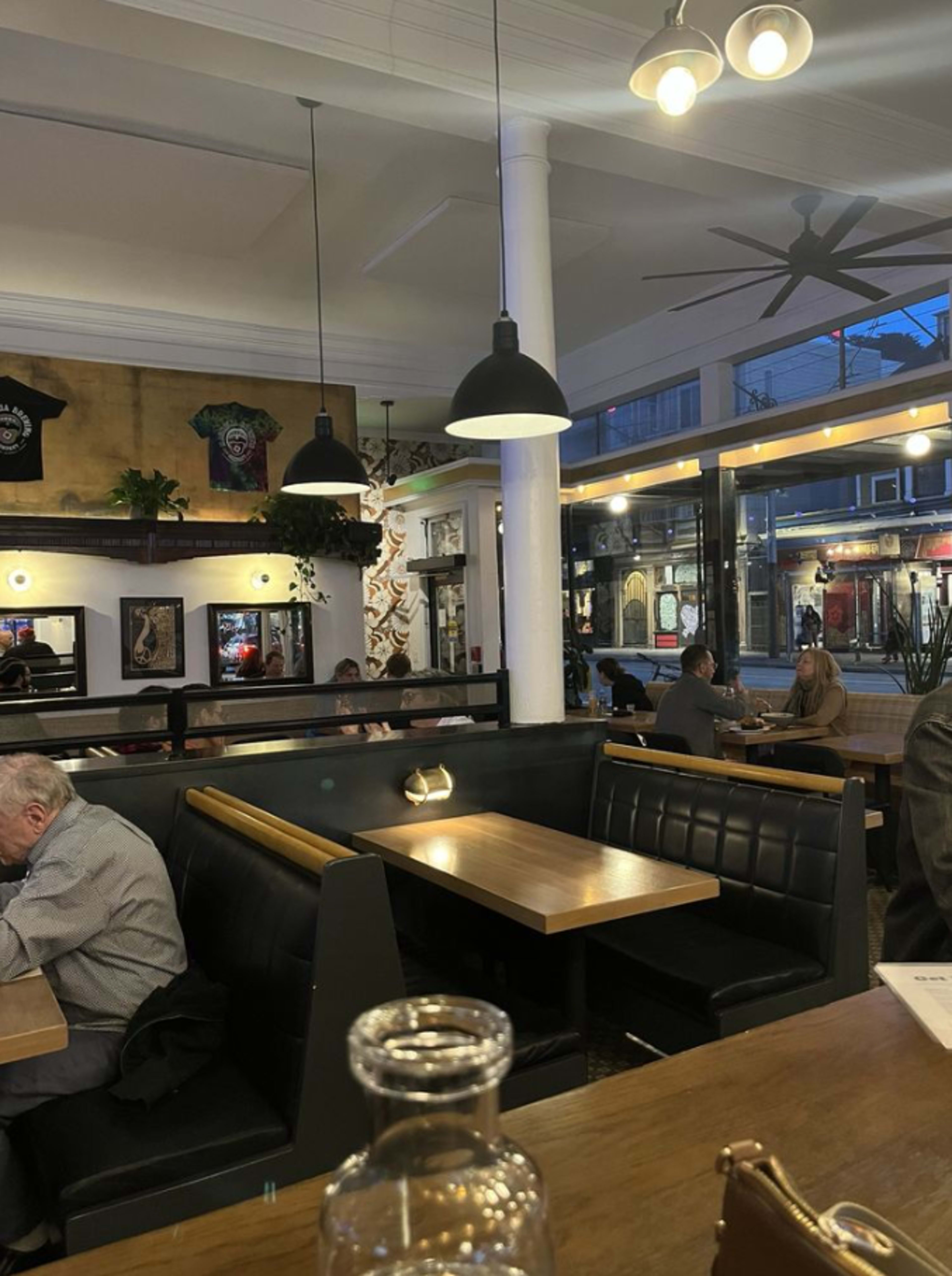 The image shows a modern restaurant interior with wooden tables, black seating, and large windows overlooking a street, while patrons dine in a relaxed atmosphere.