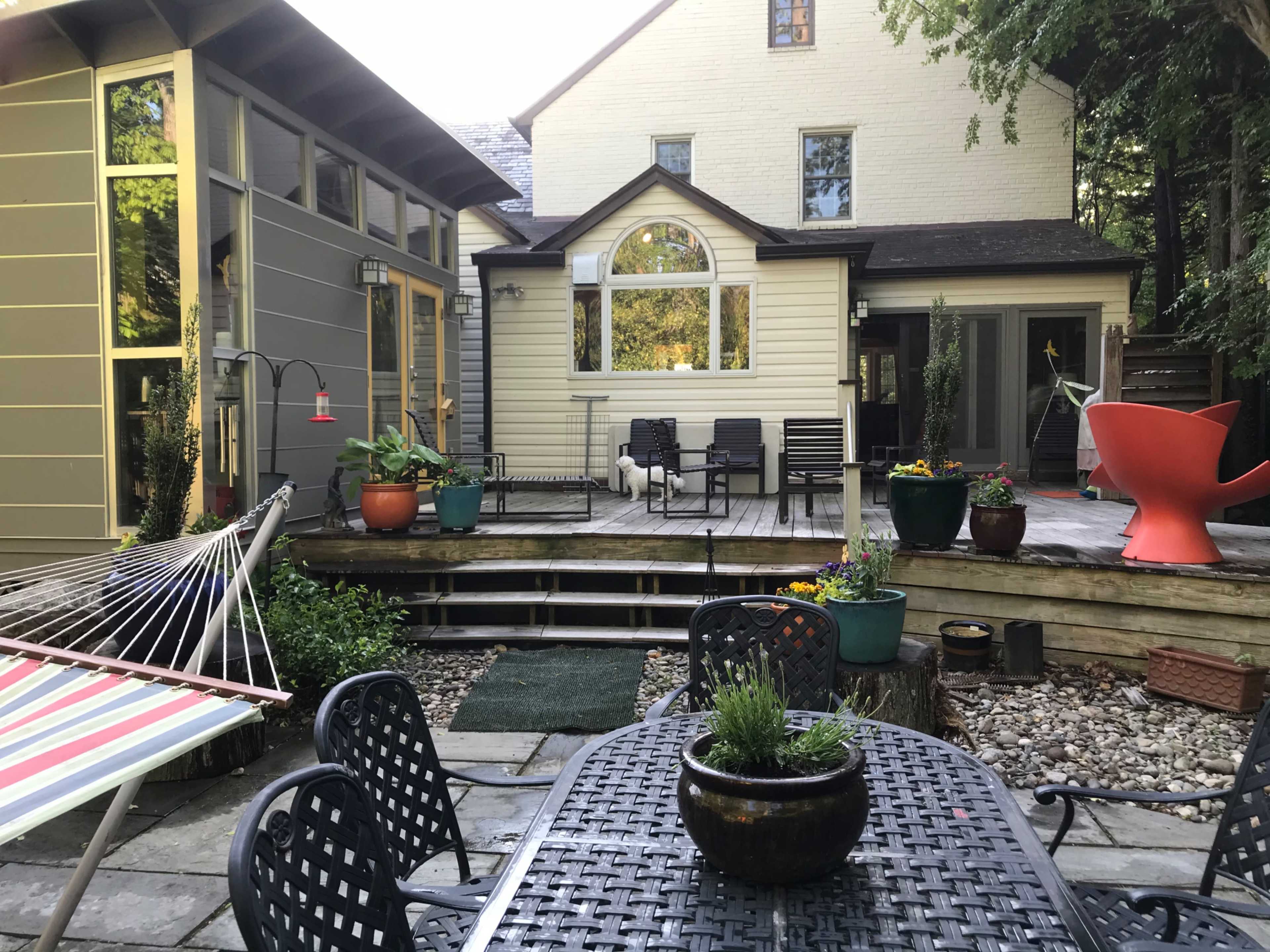 A backyard patio area with a table and chairs on a wooden deck surrounded by potted plants and a hammock.