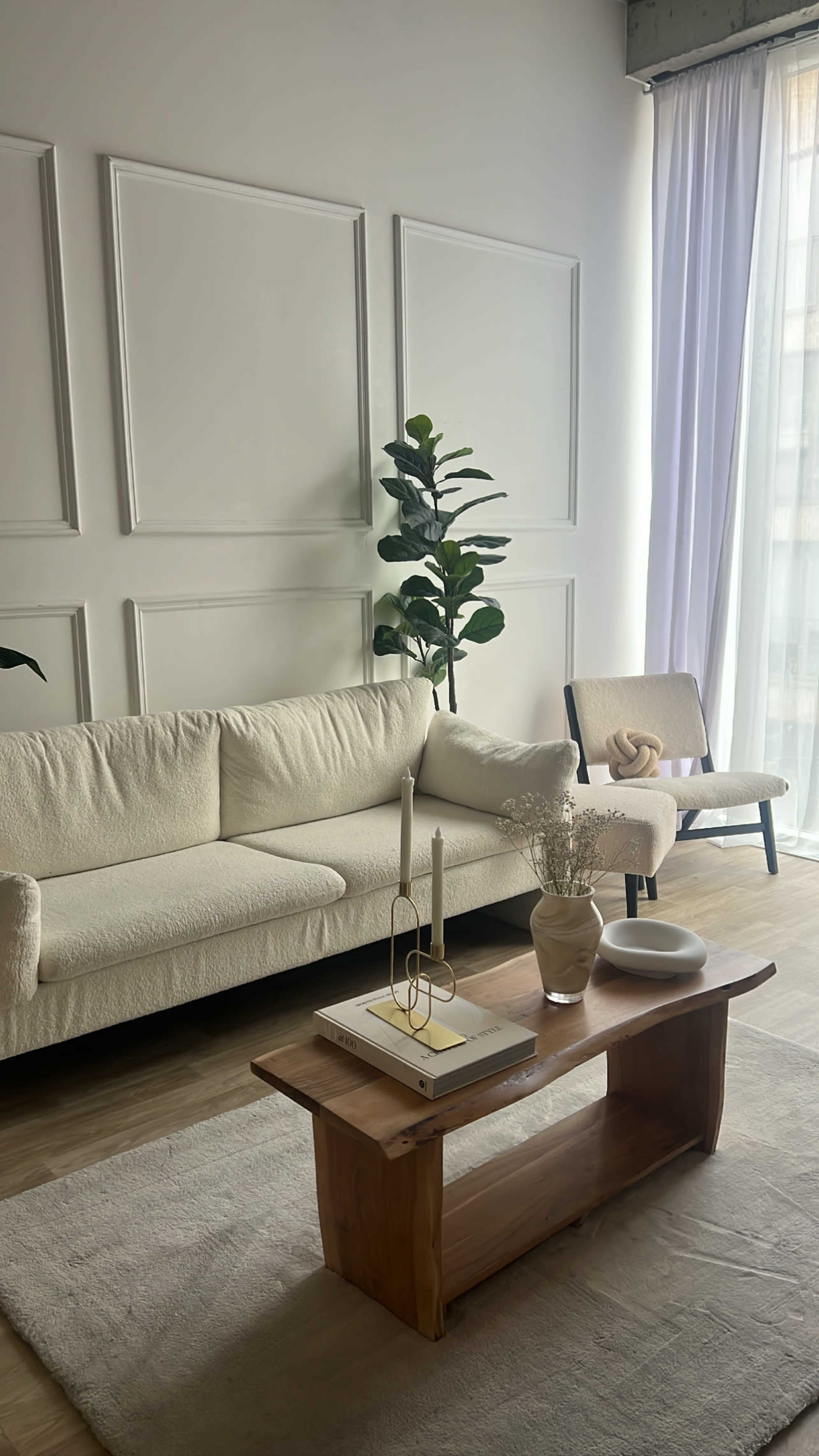 A modern living room with a white sofa, a wooden coffee table, and a tall plant beside a large window with sheer curtains.