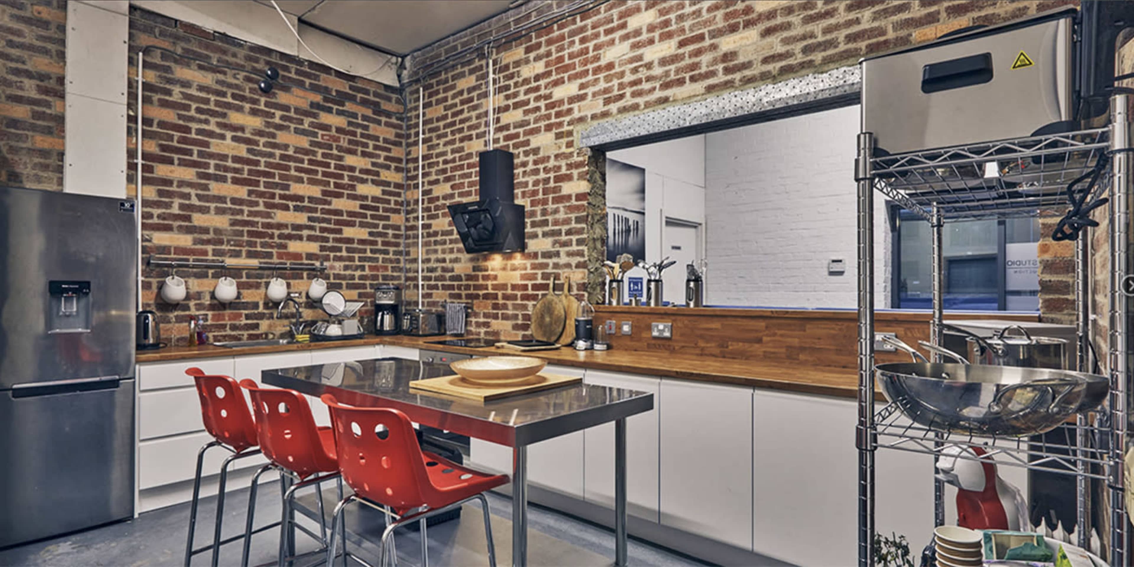 The kitchen features a brick wall, stainless steel countertops, and red plastic chairs around a rectangular table.