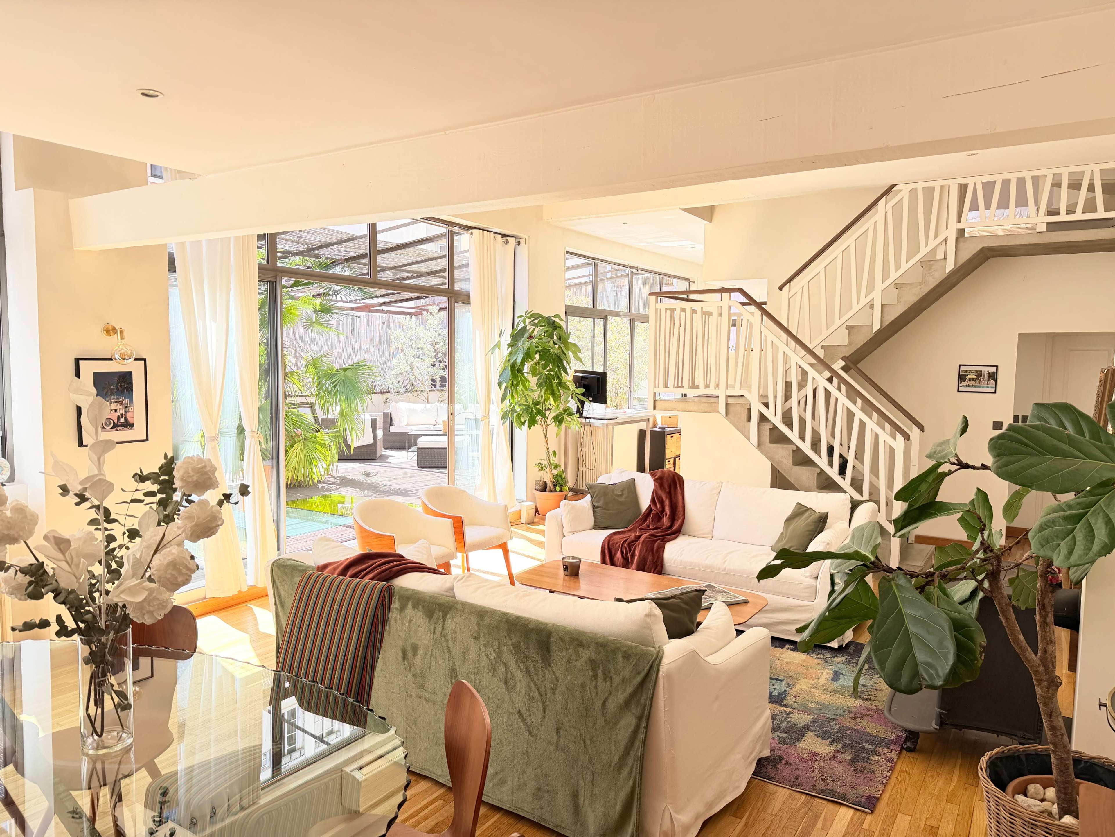 The image shows a spacious, well-lit living area featuring a mix of seating options, indoor plants, and a staircase leading to an upper level.