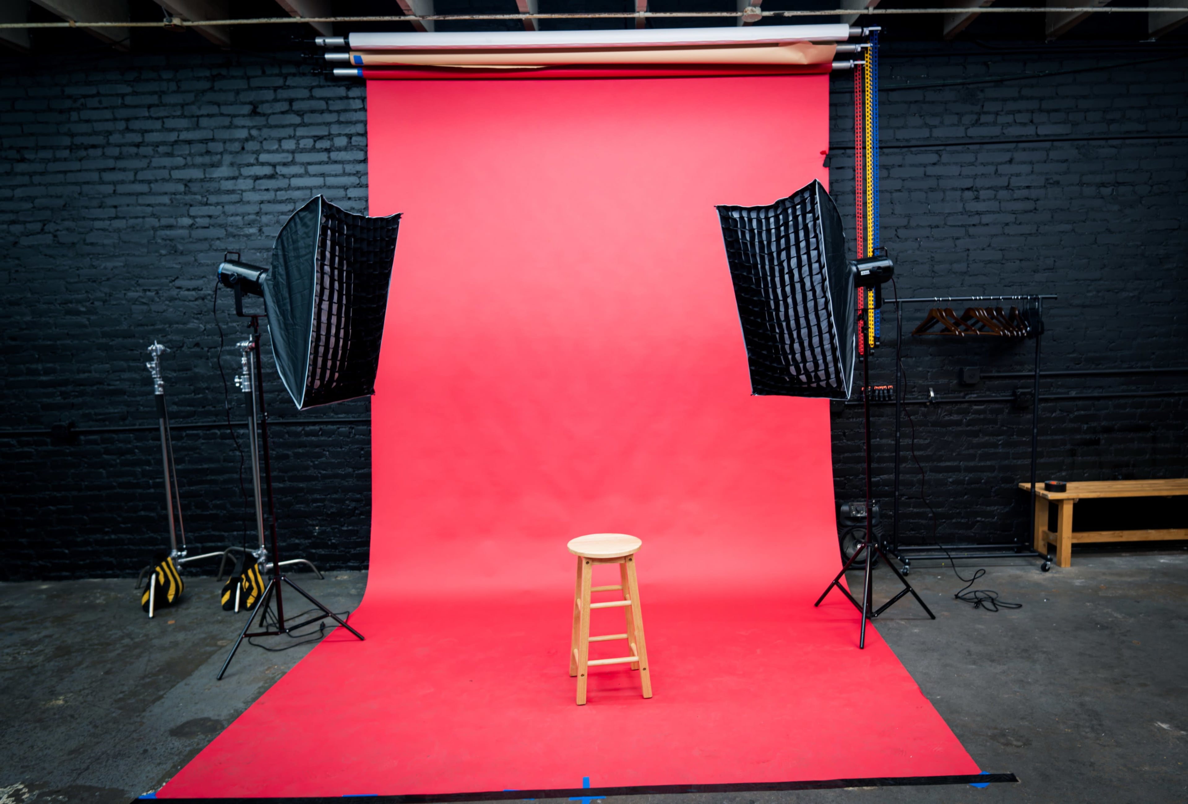 A photography studio features a red backdrop with a wooden stool in the center, flanked by two softbox lights.