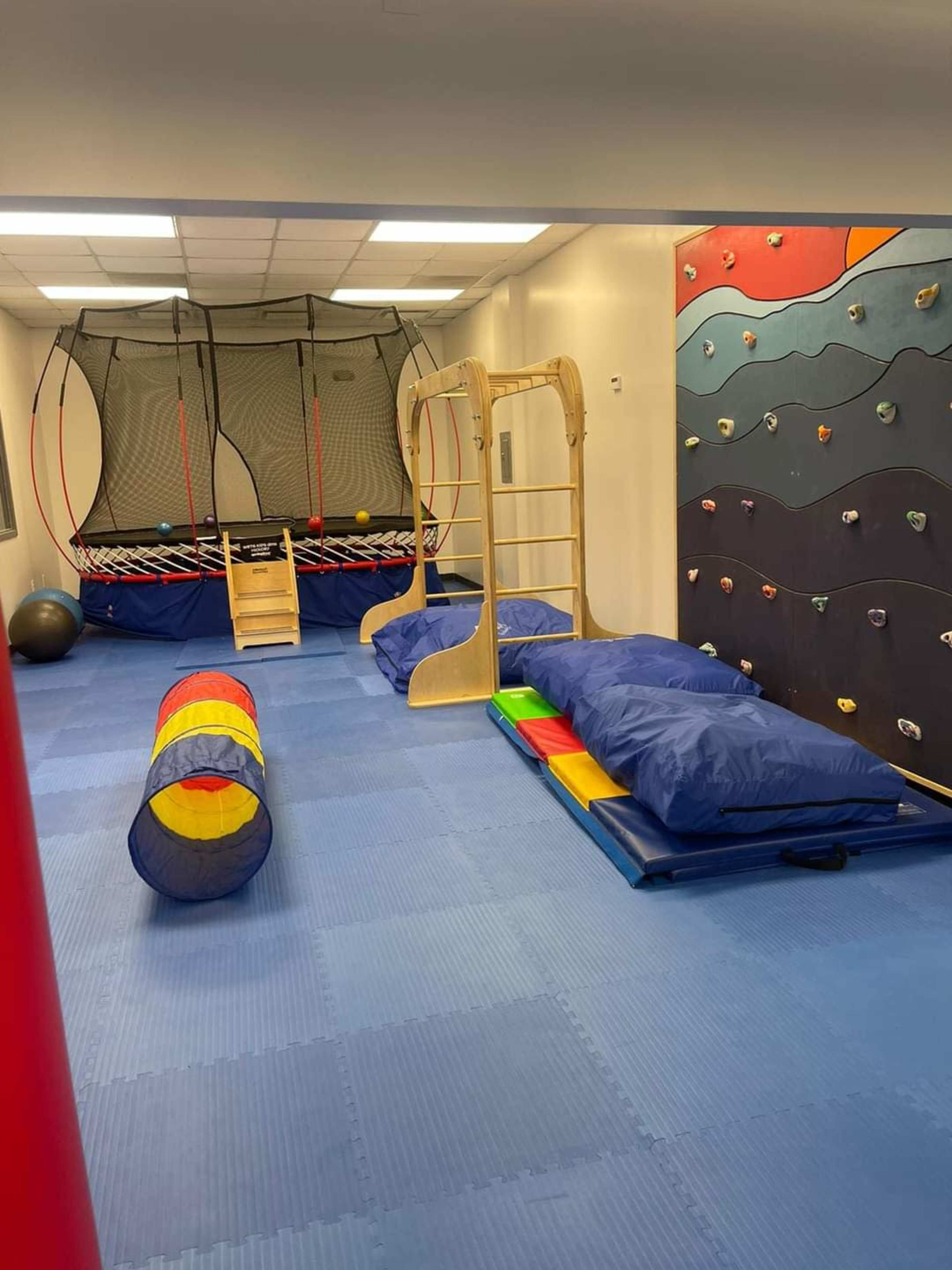 An indoor play area featuring a trampoline, climbing wall, gymnastics equipment, and colorful mats.
