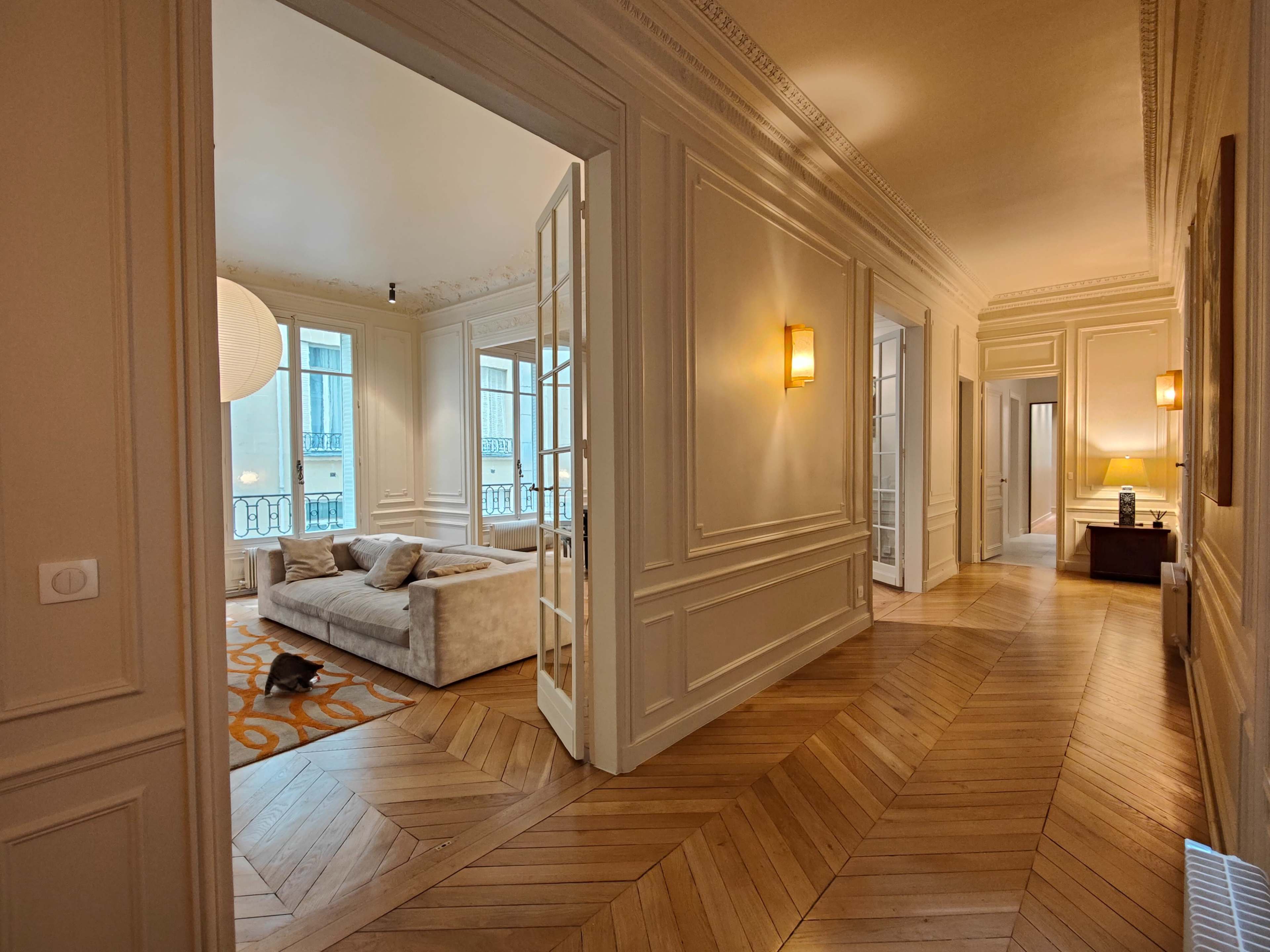 A hallway leads to a living room with a light-colored sofa, large windows, and wooden flooring.
