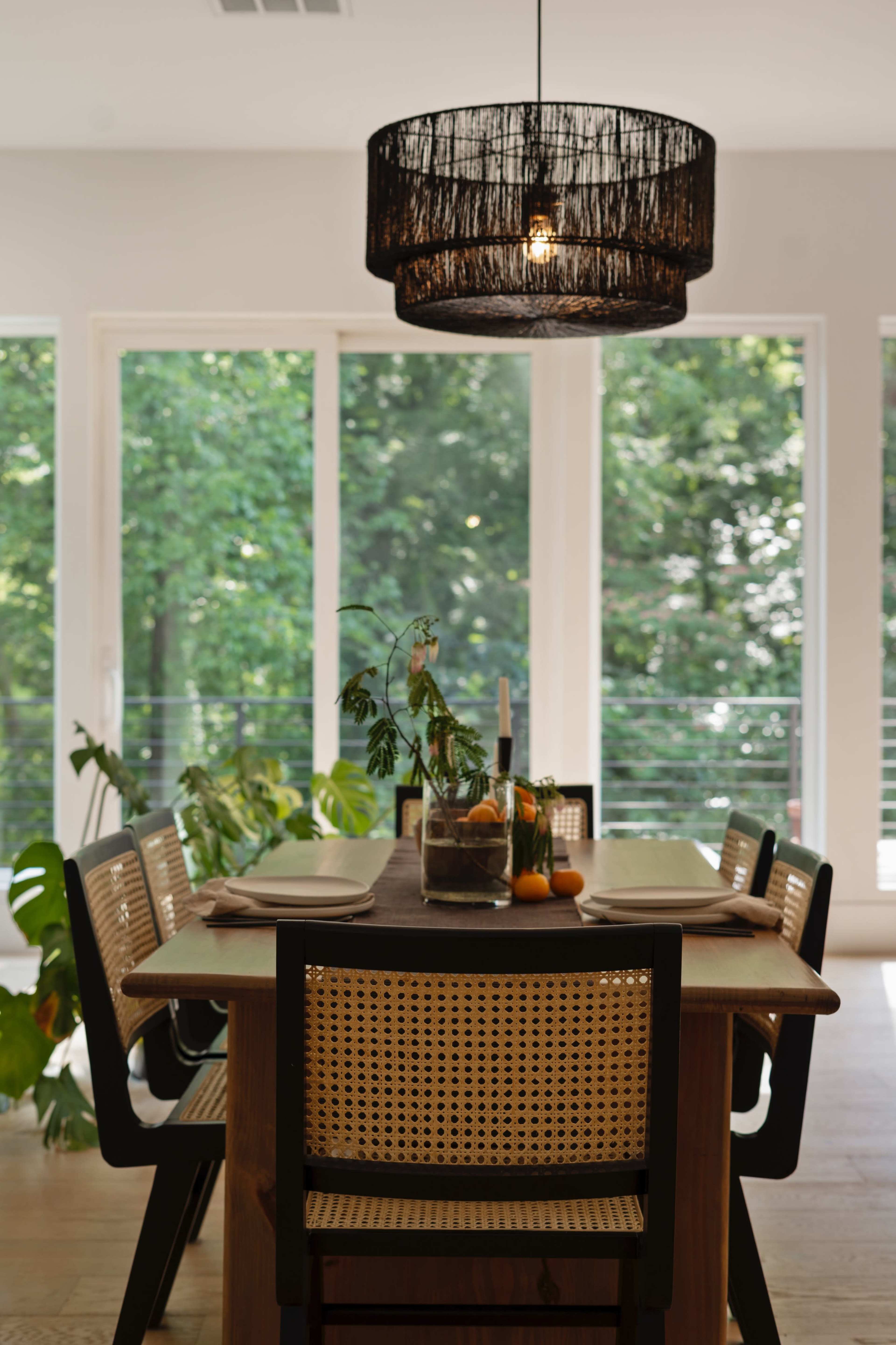 A wooden dining table is set with plates and utensils, surrounded by chairs, and illuminated by a black woven pendant light, with large windows showcasing a green, natural landscape outside.