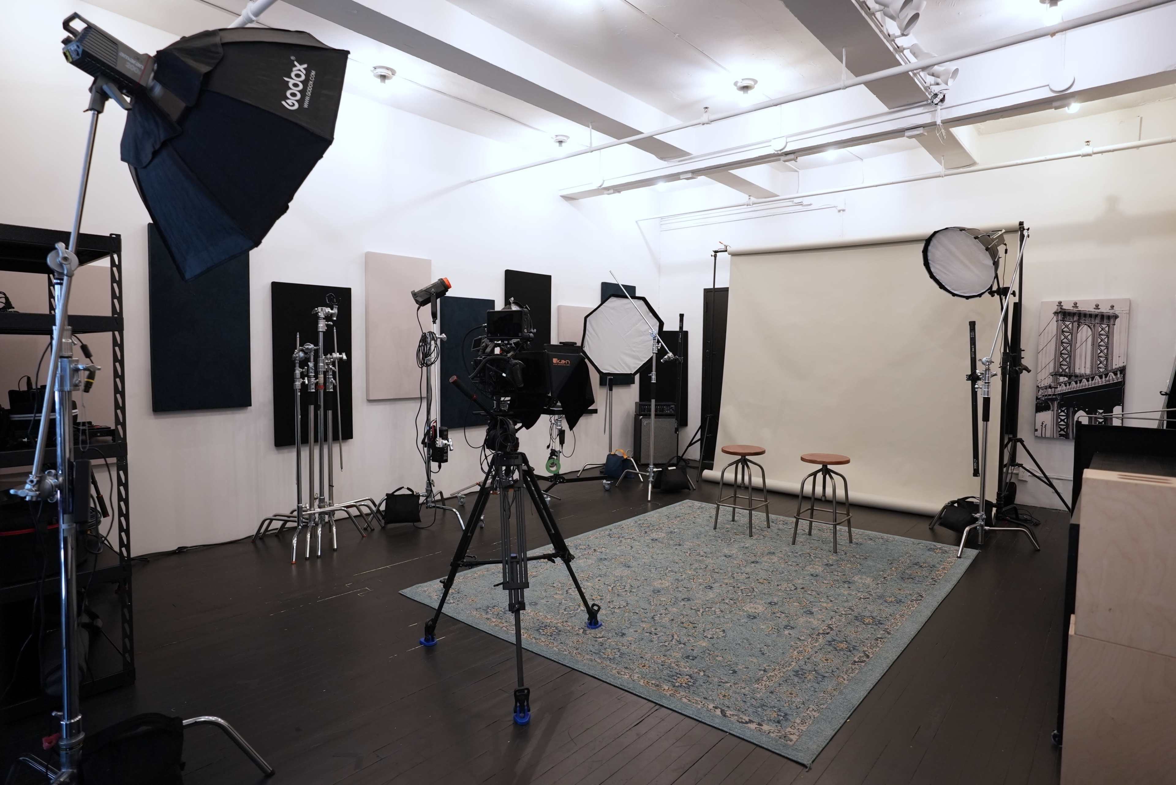 A photography studio is set up with multiple lights, cameras, and two stools against a plain backdrop on a patterned rug.