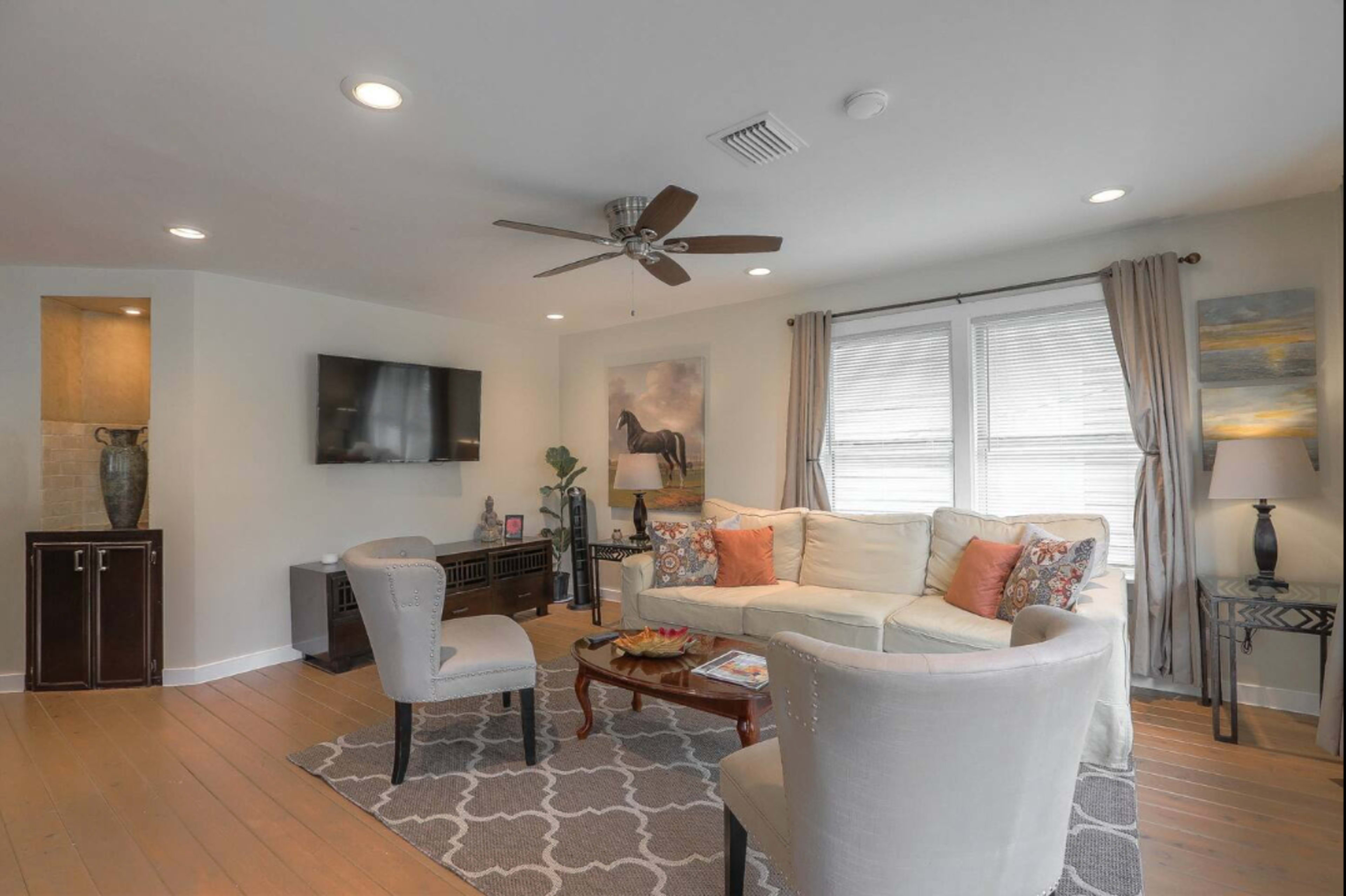A living room features a white sofa, two light-colored chairs, and a coffee table, with a large window and a television mounted on the wall.
