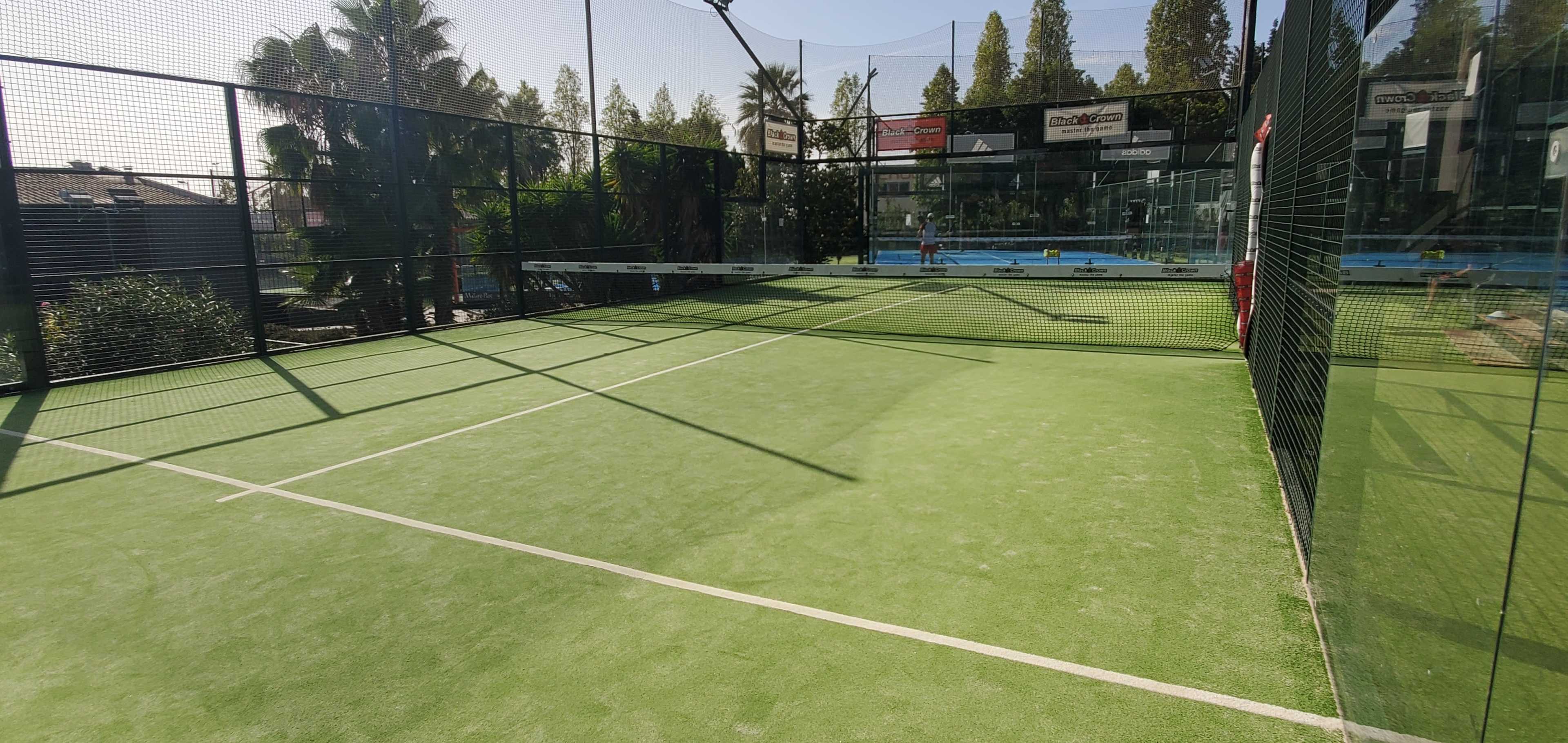 A brightly lit padel court with green artificial turf, surrounded by high fences and palm trees in the background.