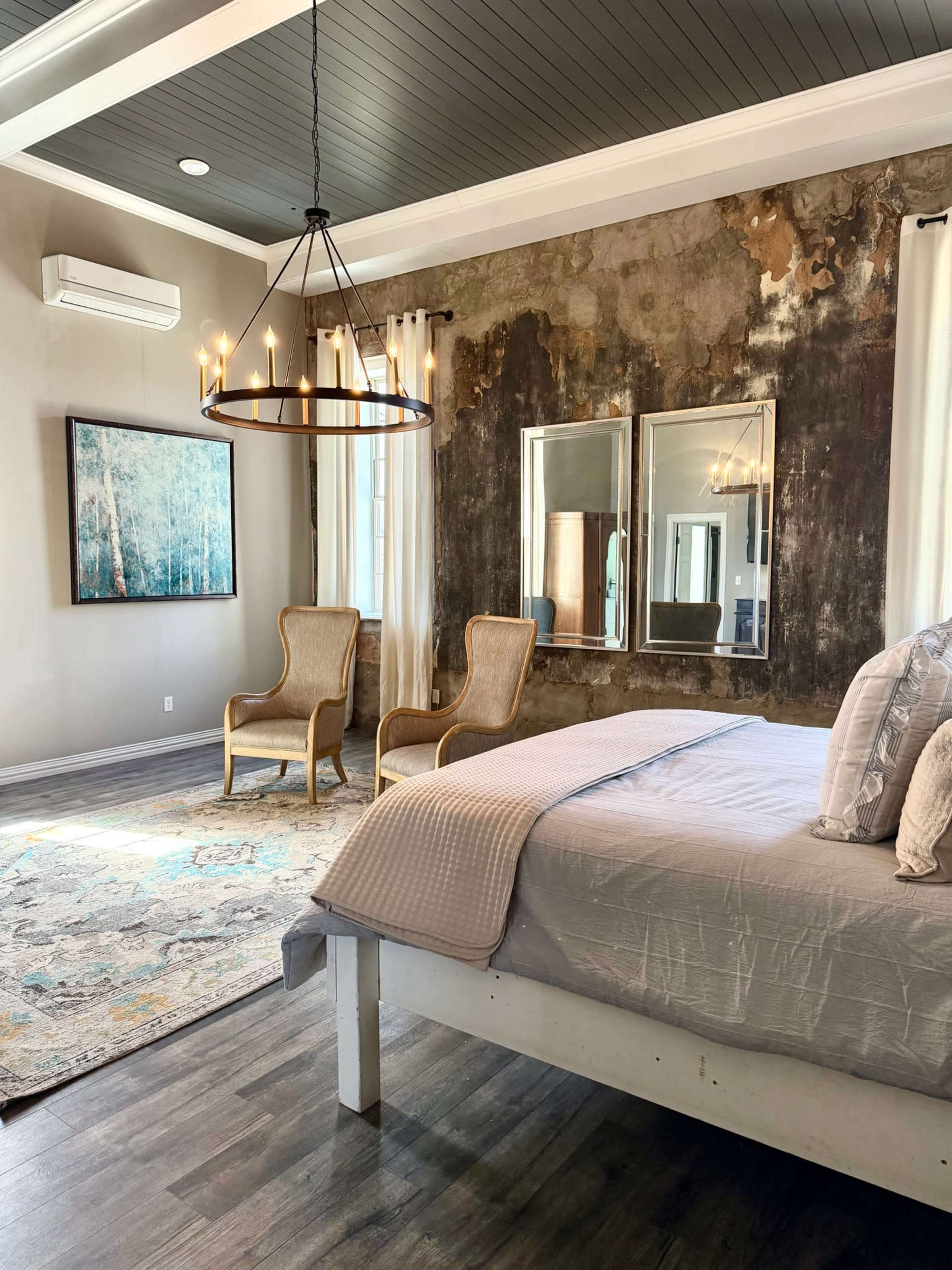 The image shows a modern bedroom with a bed, two armchairs, a large area rug, and a chandelier, featuring a textured wall and large mirrors.