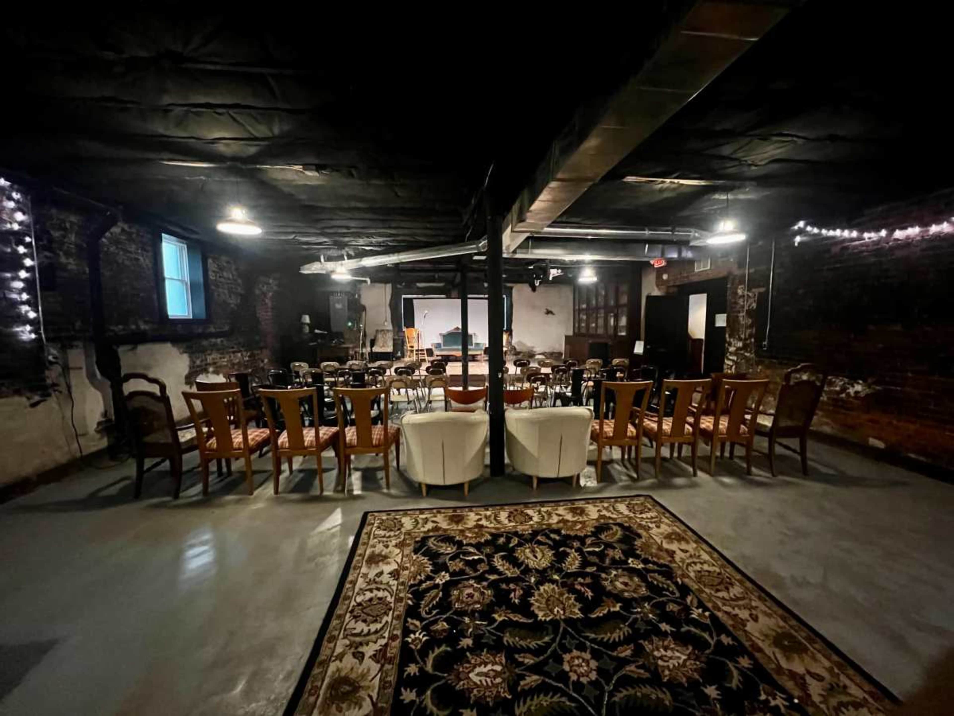 The image shows a dimly lit basement space furnished with various chairs arranged in a semicircle around a central area, with a large white screen and a patterned rug in the foreground.