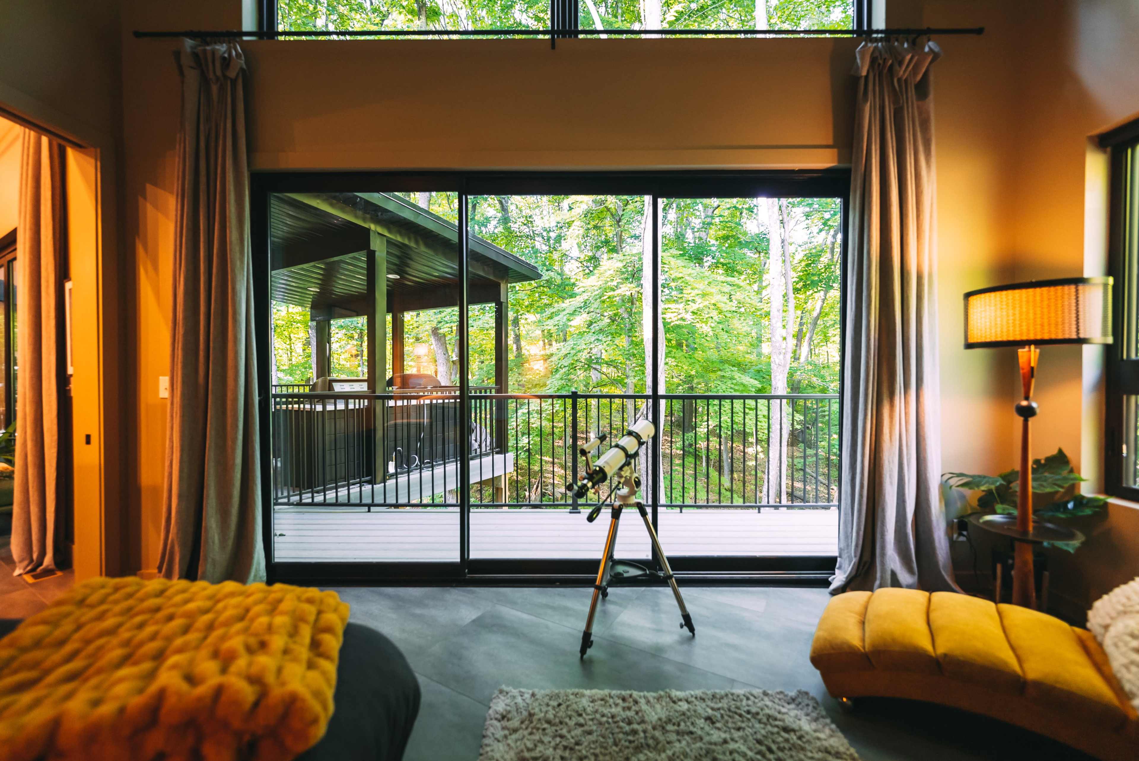A cozy room features a telescope positioned near large glass doors that open to a wooded balcony.