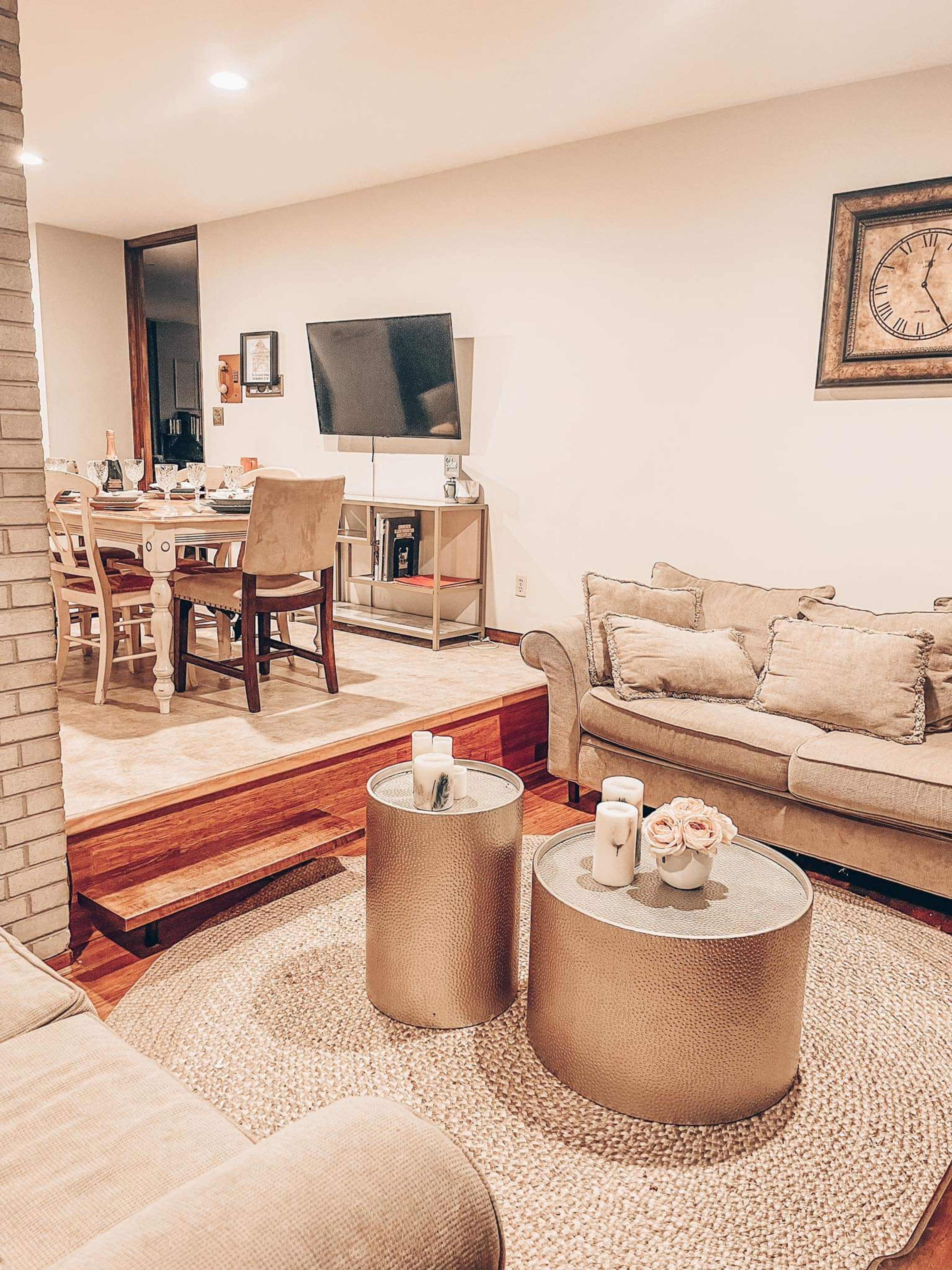 A modern living room features a beige sofa, two gold side tables, and a television mounted on the wall, leading to a dining area with a table and chairs.
