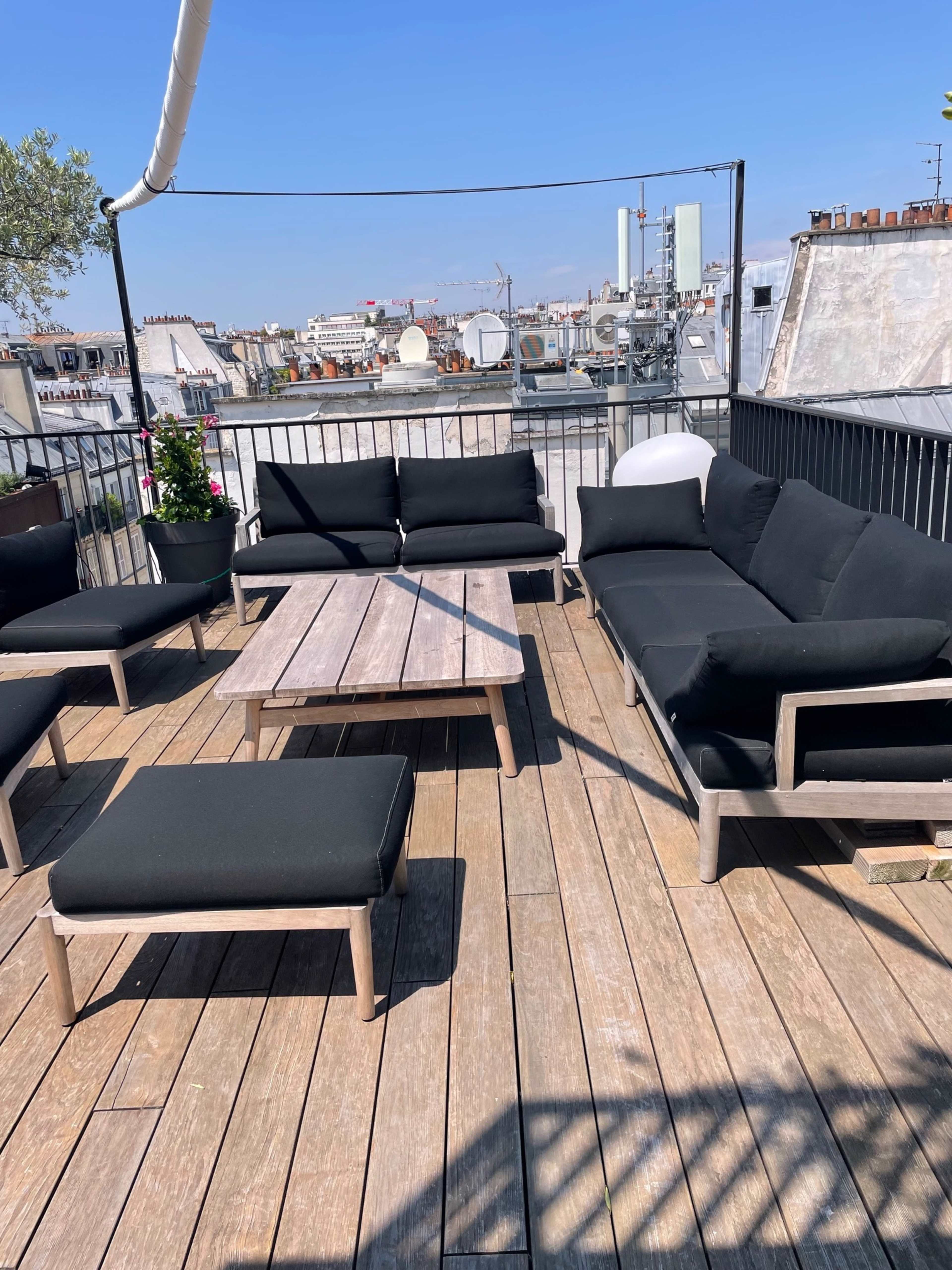 The image shows a rooftop terrace with wooden decking, featuring black outdoor couches and a coffee table, set against a clear blue sky.