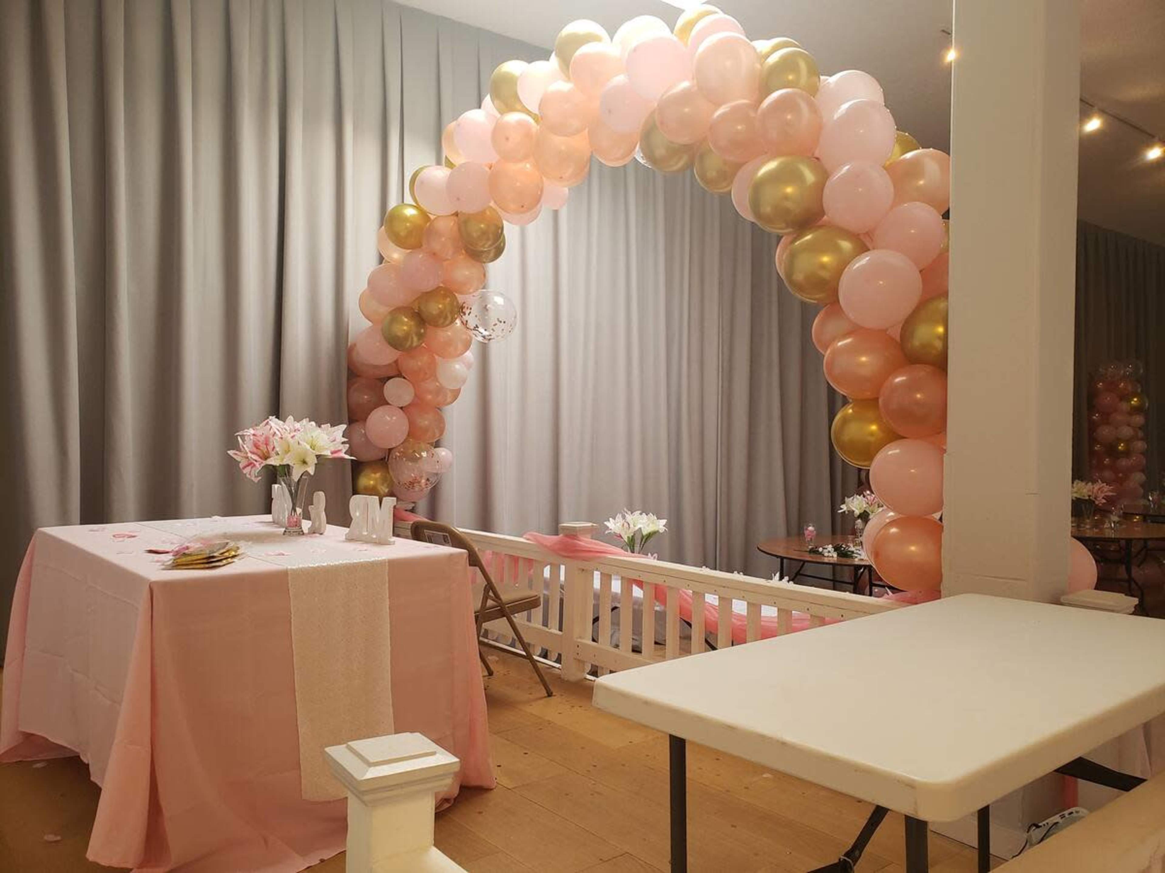 A festive space is decorated with an arch of pink, gold, and white balloons and a table set with flowers and tableware.