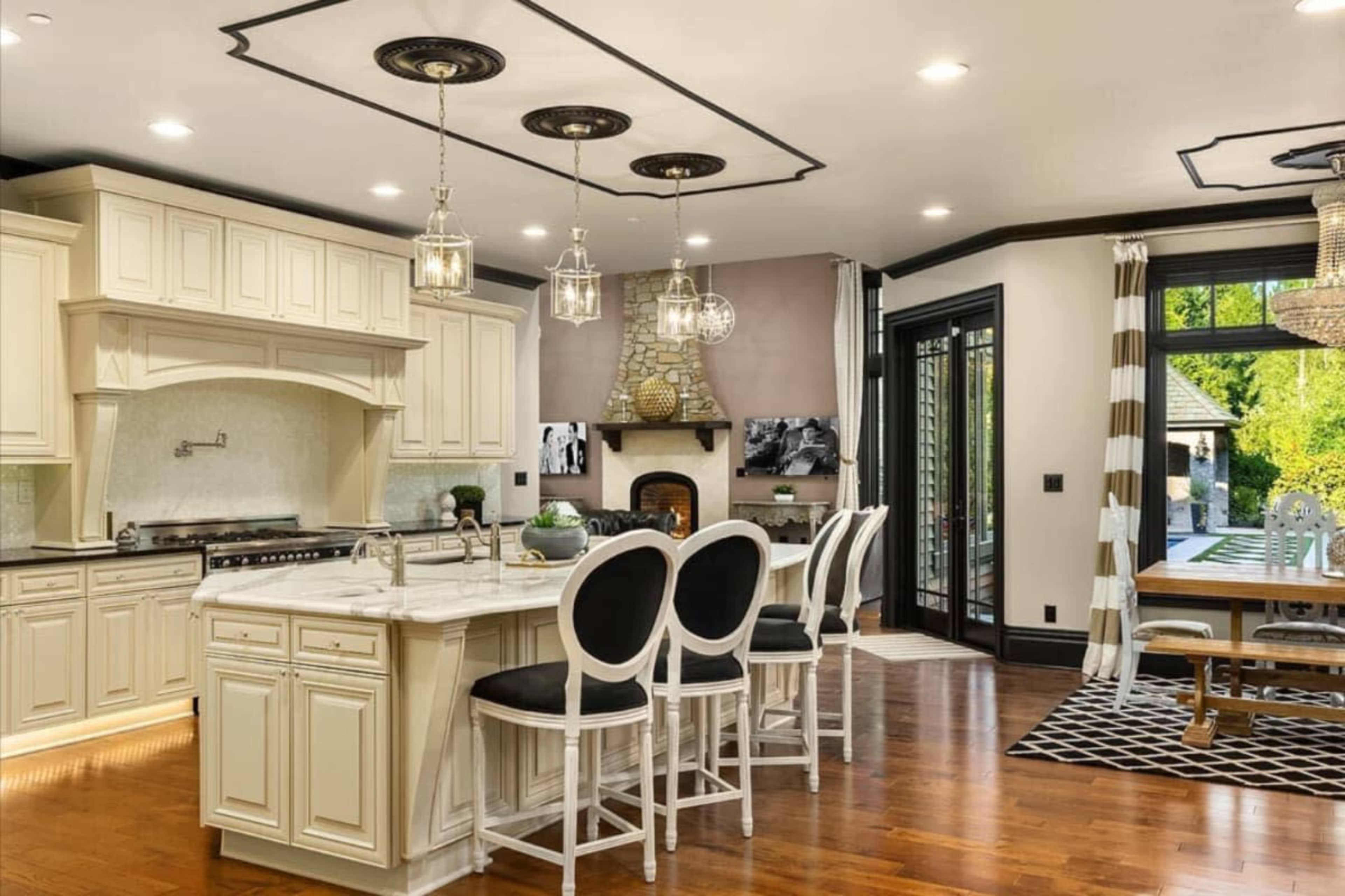 A modern kitchen features white cabinetry, a large marble island with black stools, and a dining area with a black and white rug.