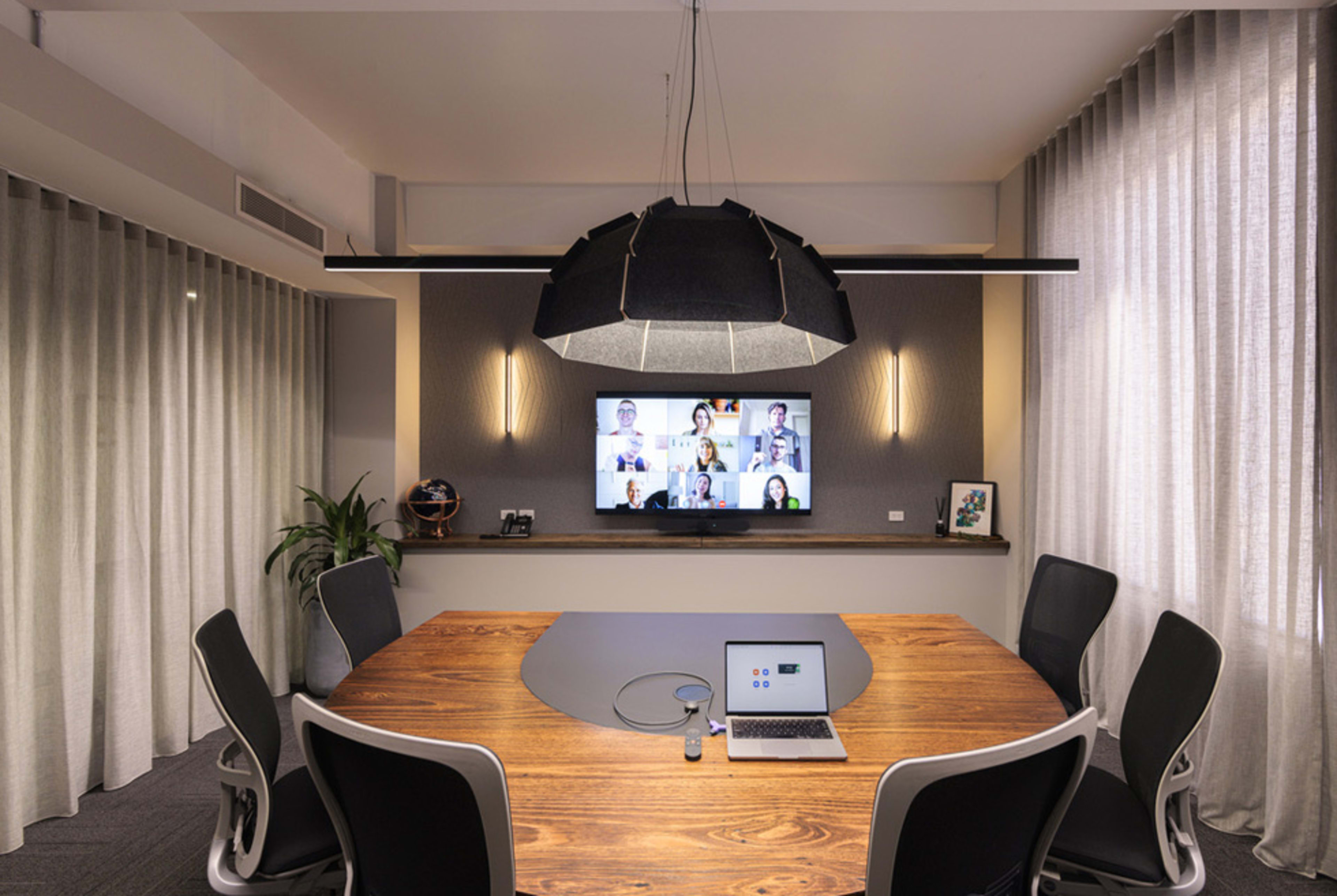 A modern conference room features a large wooden table at the center, surrounded by ergonomic chairs, with a screen on the wall displaying a video call.