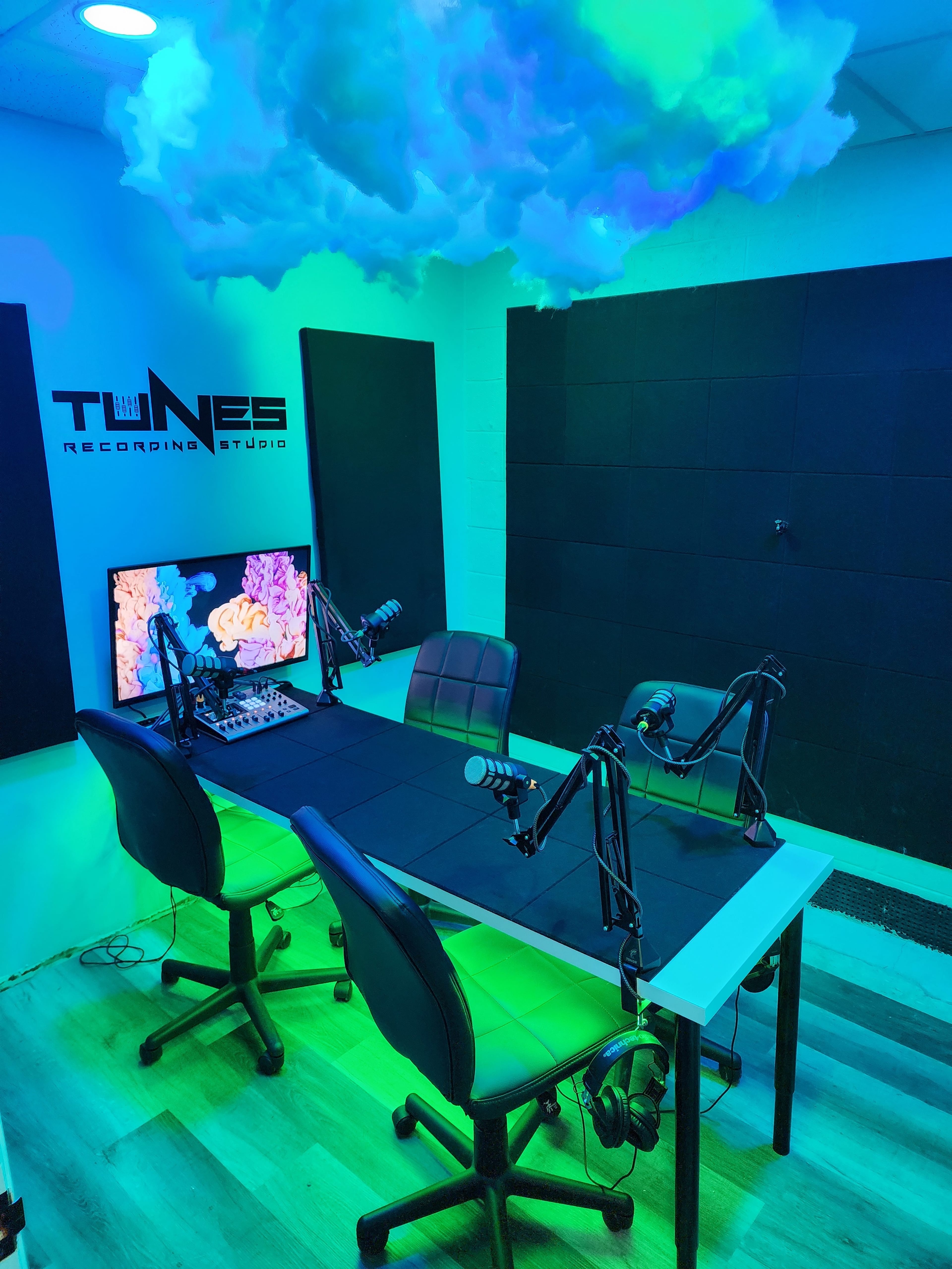 A recording studio with two black chairs, a table equipped with microphones, a computer screen displaying visual elements, and decorative cloud ceiling features.