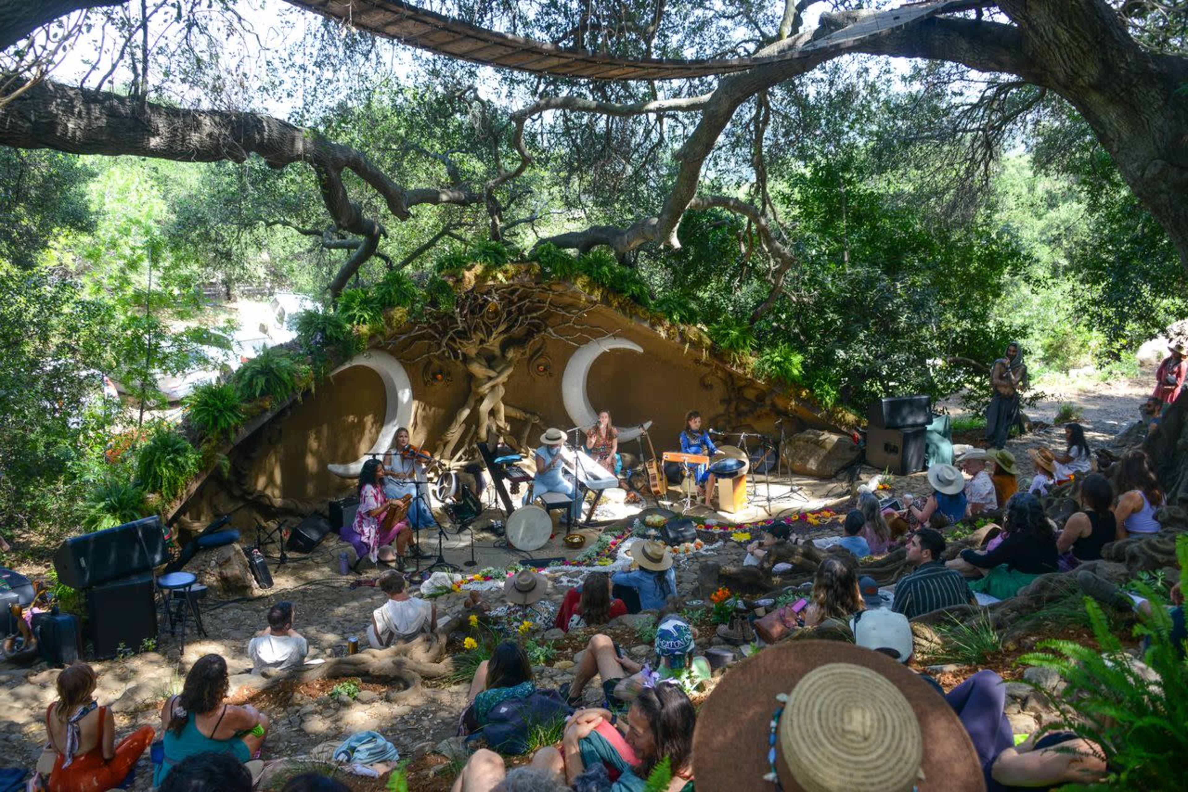 A diverse group of people is gathered in a natural setting, watching a band perform in front of an intricately designed stage made from natural materials.