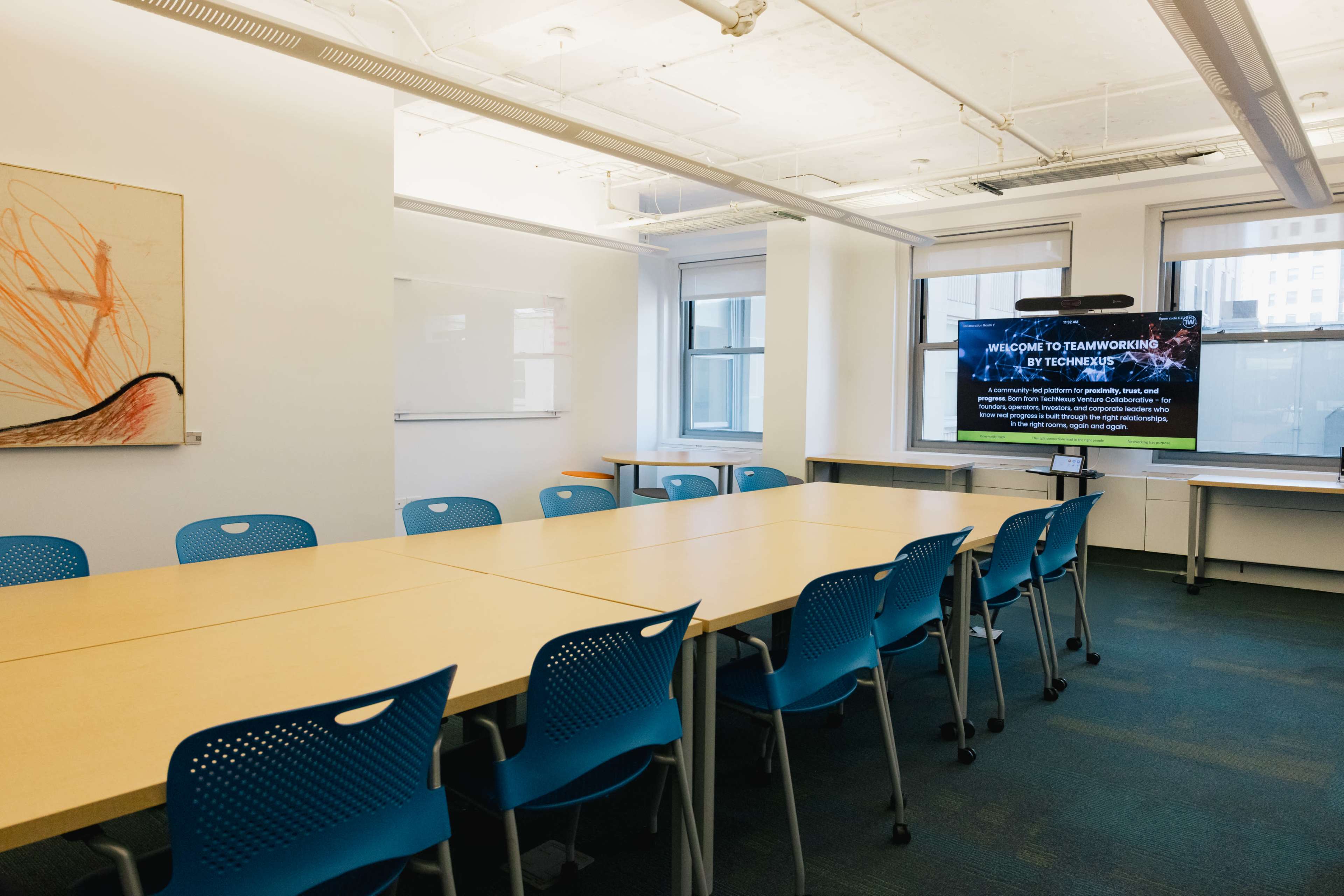The image shows a modern meeting room with a long wooden table surrounded by blue chairs, and a screen displaying a presentation in the background.