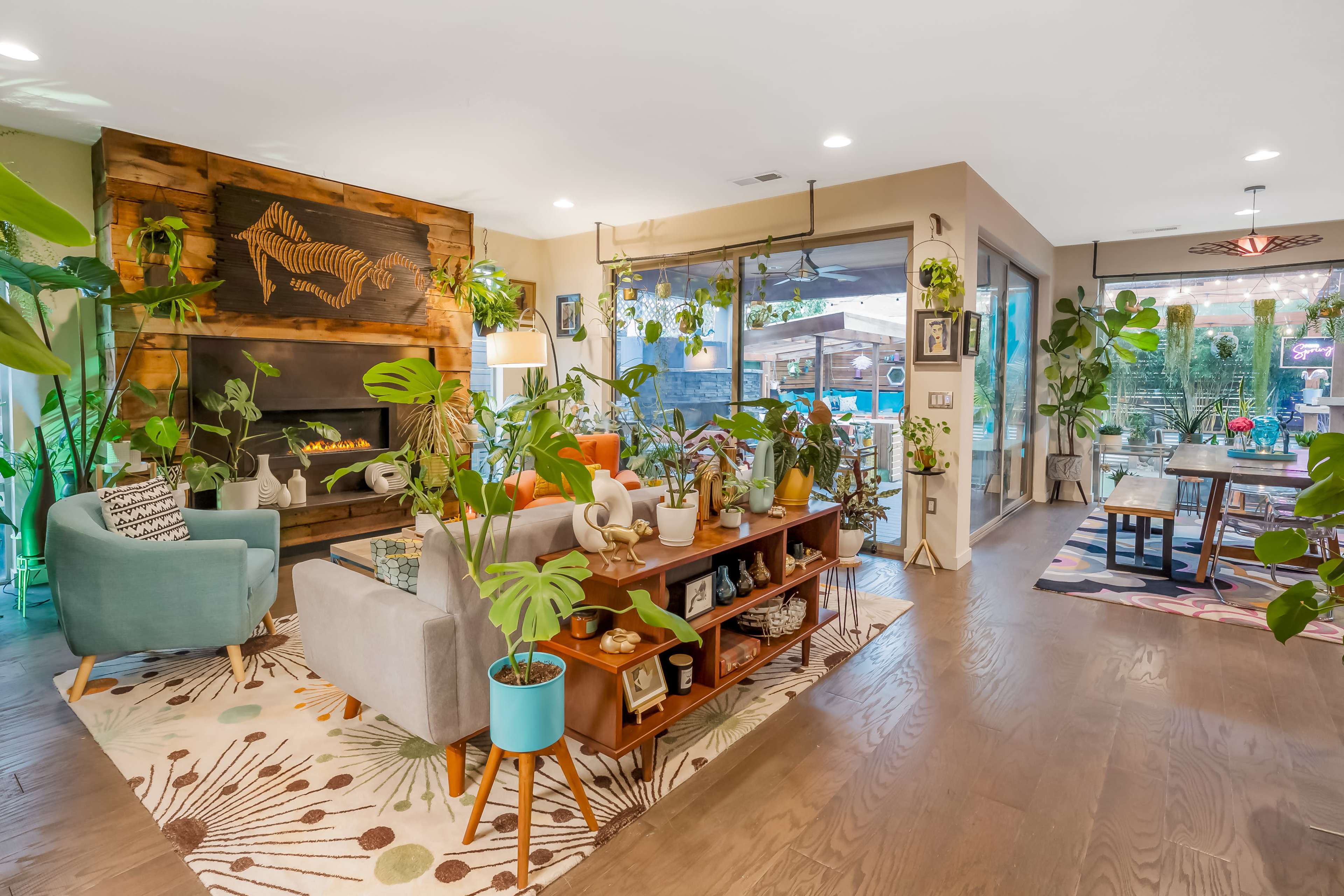 A modern living room decorated with plants, a wooden fireplace, and a cozy seating arrangement that opens up to an outdoor space.