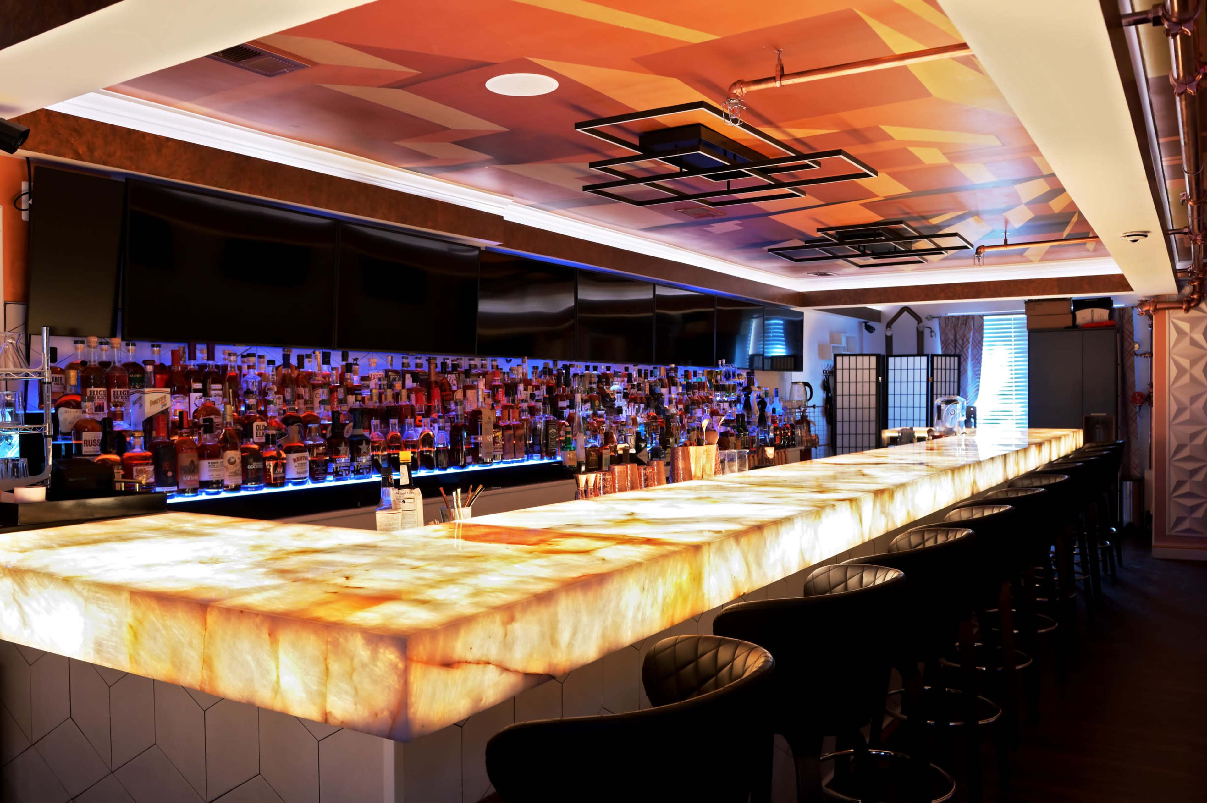 The image shows a modern bar with an illuminated onyx countertop and a back wall lined with various bottles of alcohol.