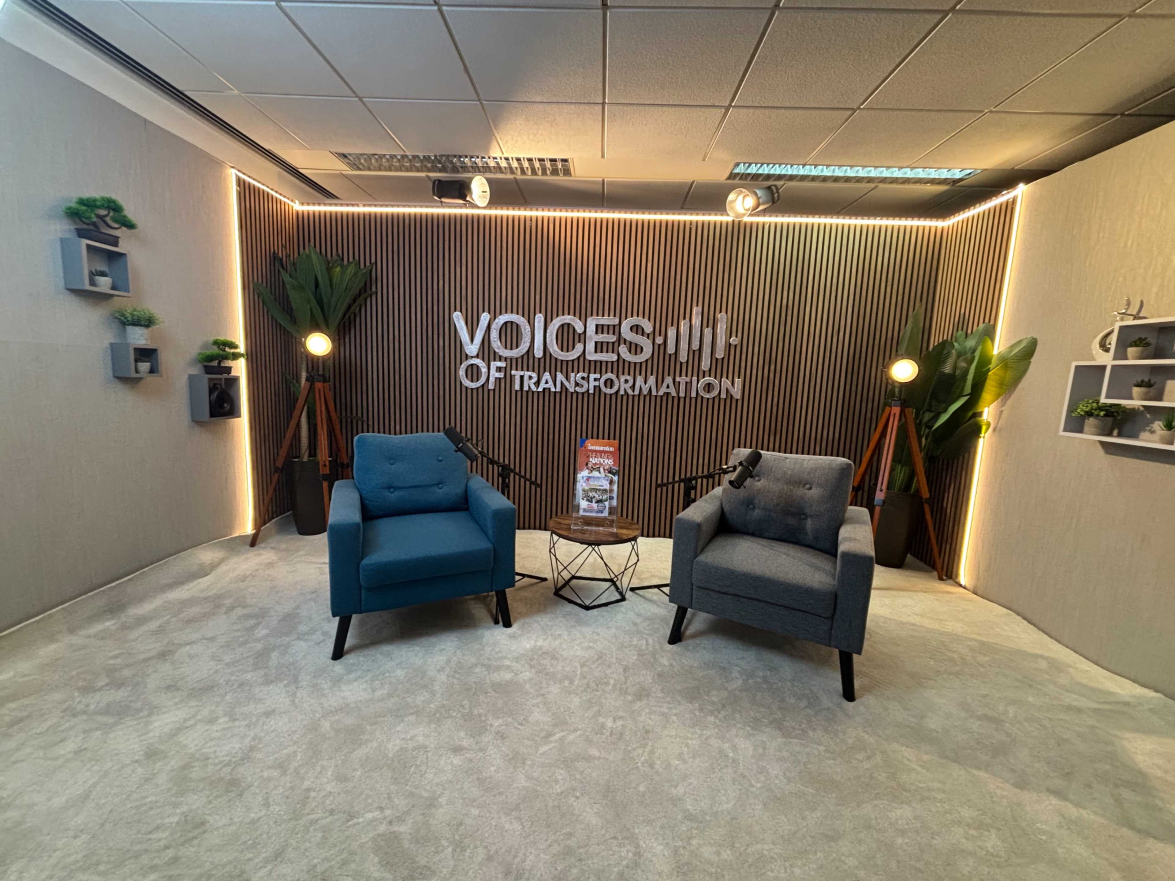 The image shows a modern, well-lit studio setup featuring two armchairs, a small table, and backdrop signage reading "Voices of Transformation."