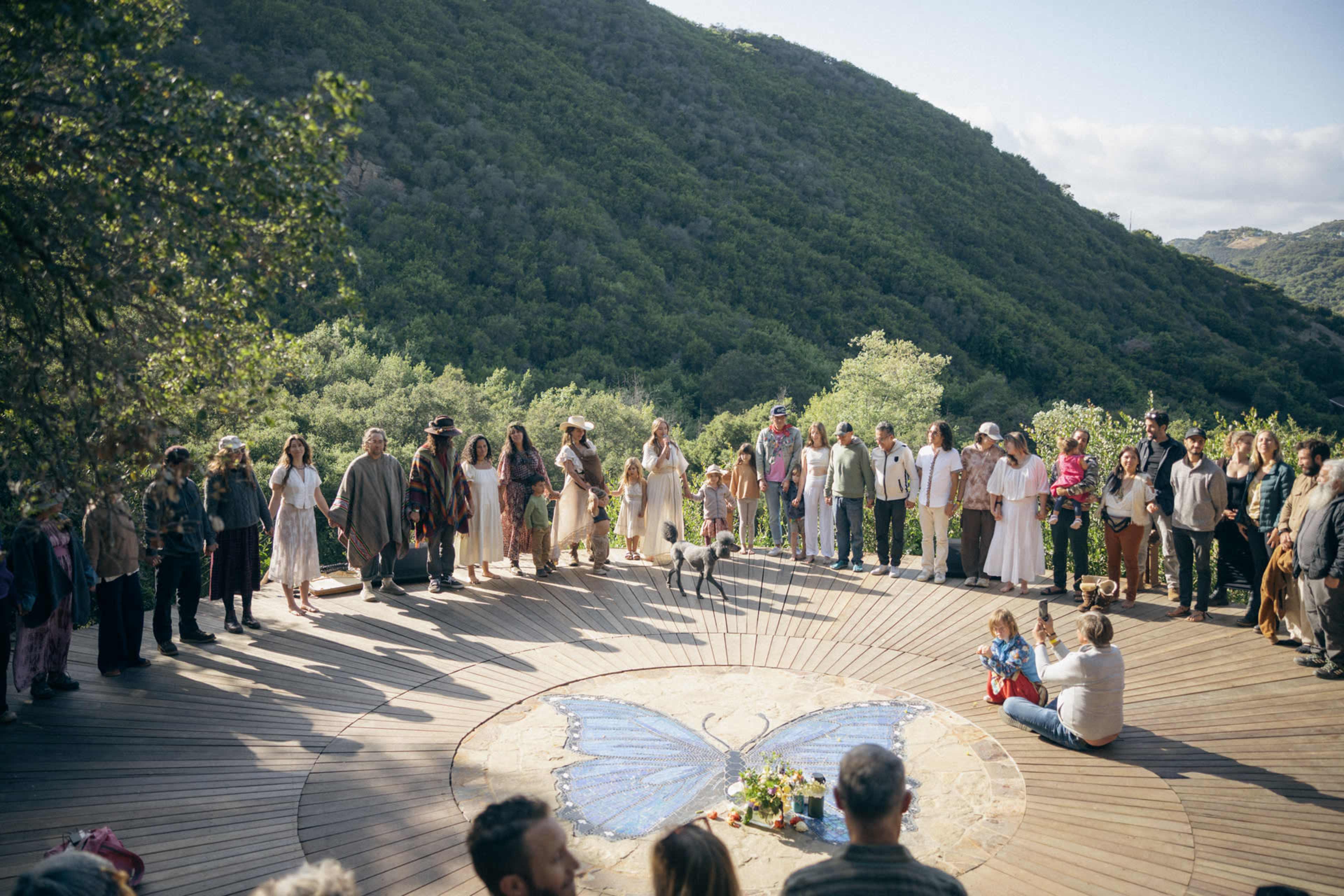 A diverse group of people stands in a circle around a butterfly mosaic on a wooden deck, set against a backdrop of lush green hills.
