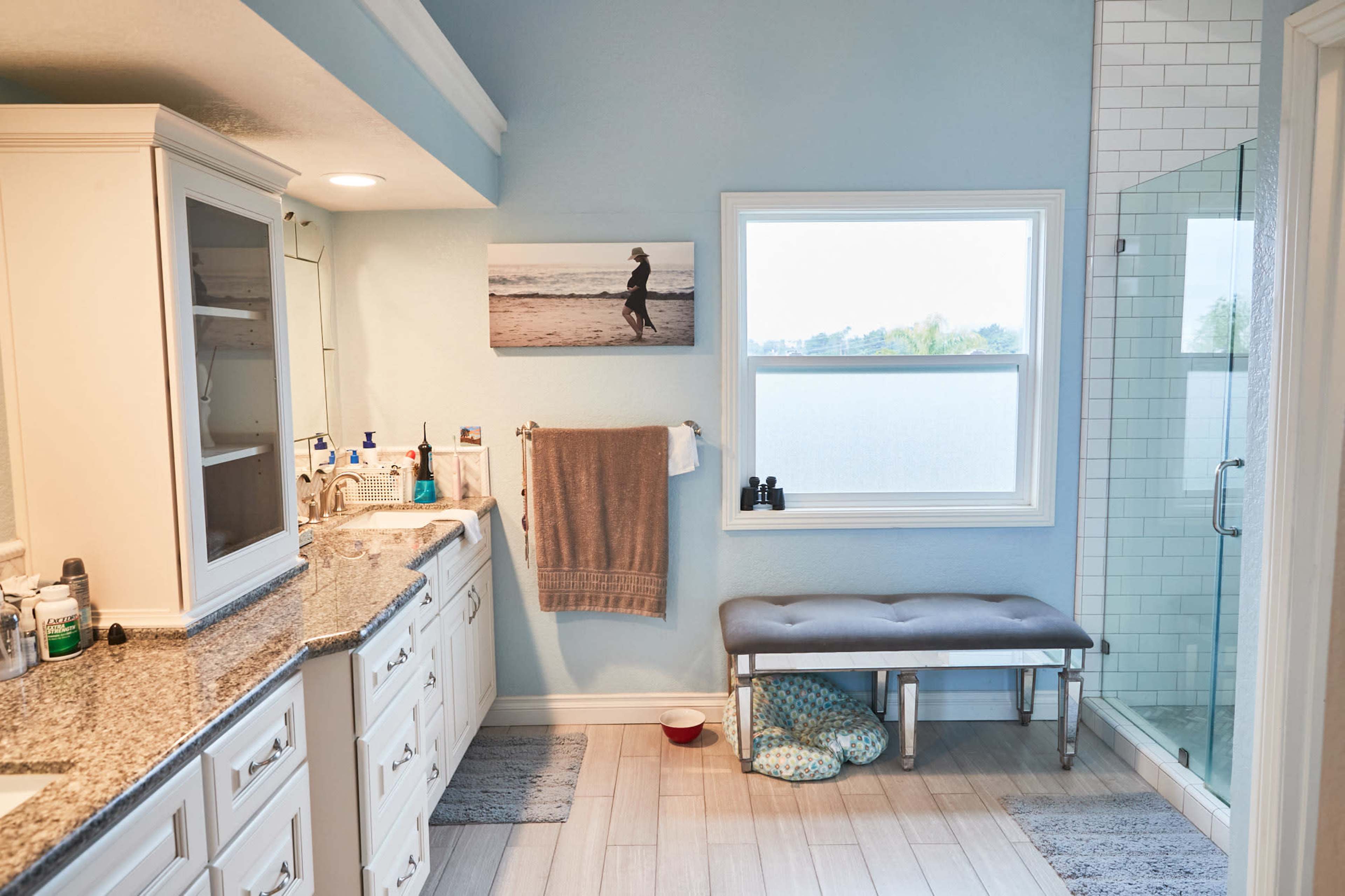 A bathroom features a double sink vanity with granite countertops, a large window overlooking a serene view, a shower enclosure, and a bench near the floor.
