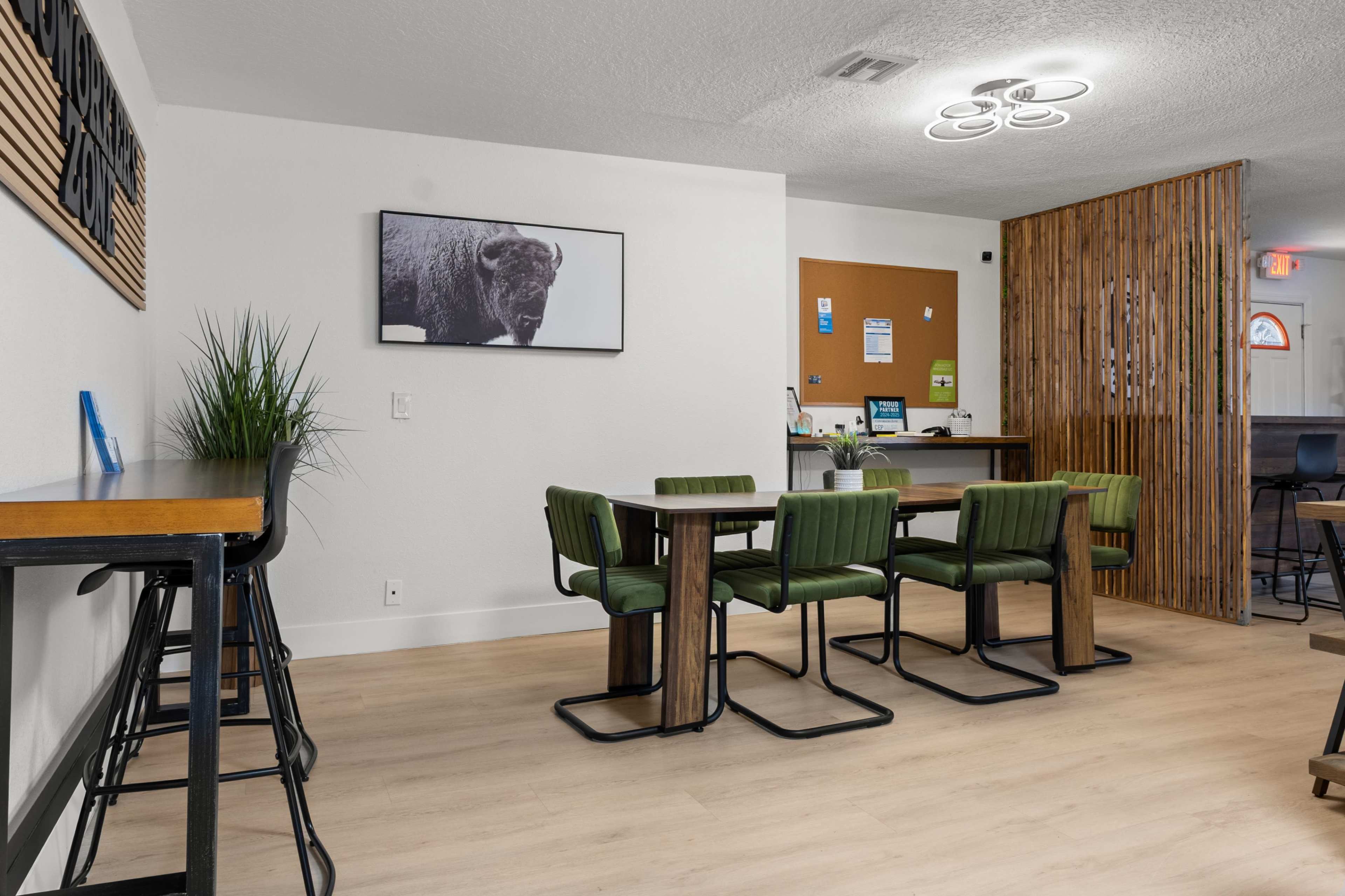 The image shows a modern communal workspace with green chairs around tables, a piece of wall art featuring a bison, and a wood-paneled reception area in the background.