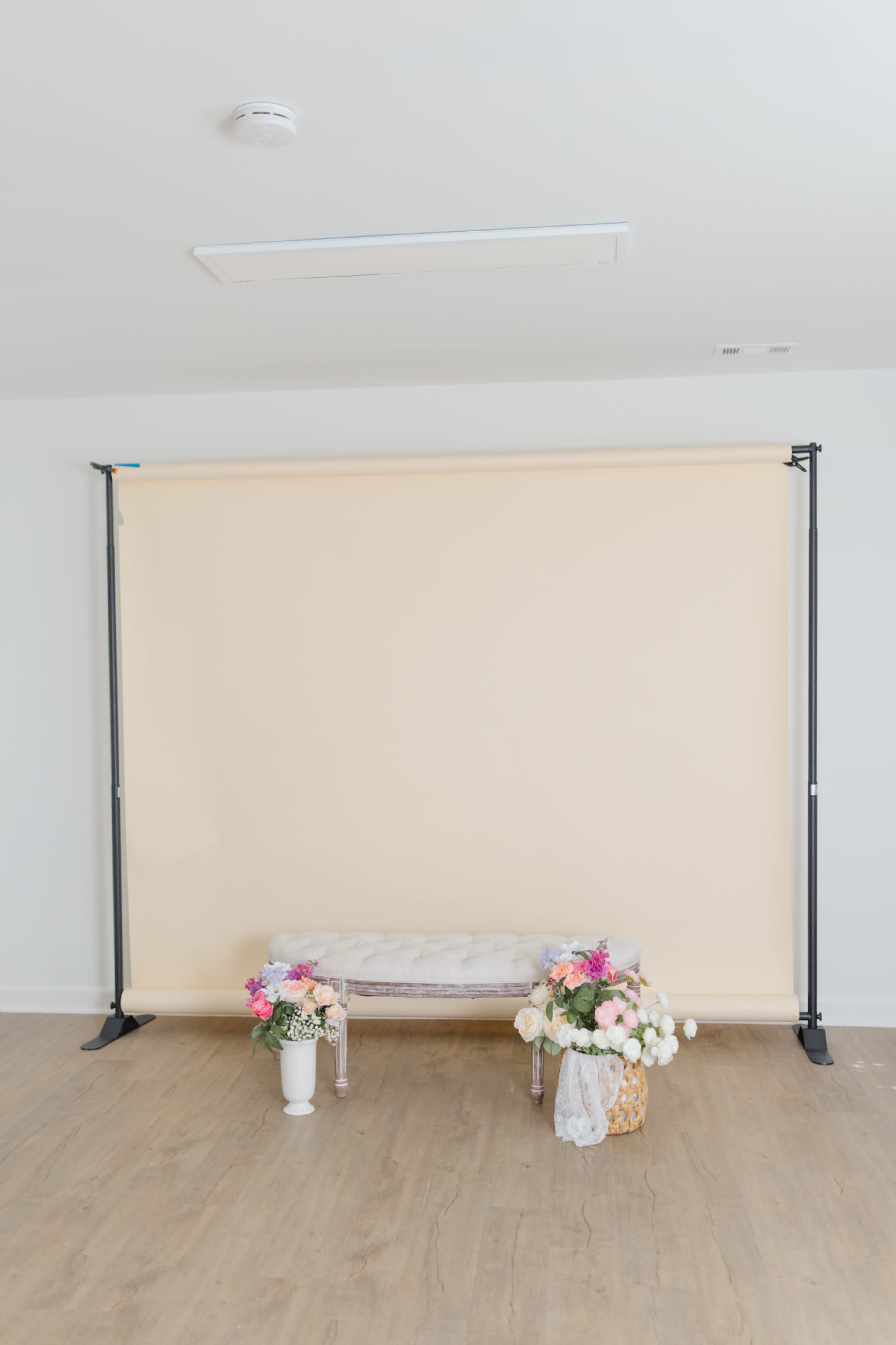 The image shows a light beige backdrop on a frame, with a decorative bench and two flower arrangements placed in front of it.